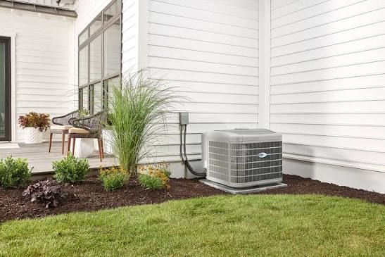 Outdoor air conditioner beside a house wall, with mulch beds, grass, and potted plants in the yard
