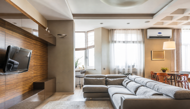 Modern living room with gray sectional sofa, wall-mounted TV, wooden accent wall, and large bright windows