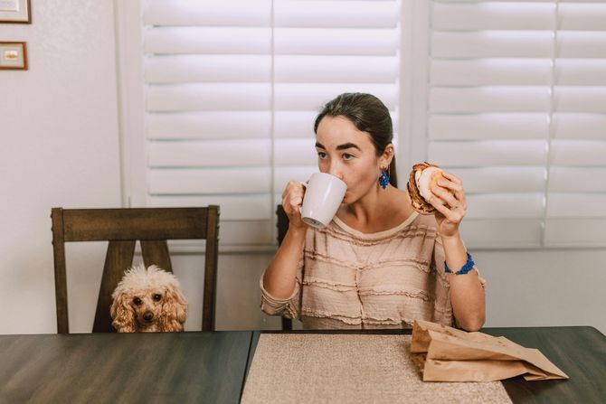 Woman at a dining table drinking from a mug and holding a bagel, with a dog peeking from a chair nearby.