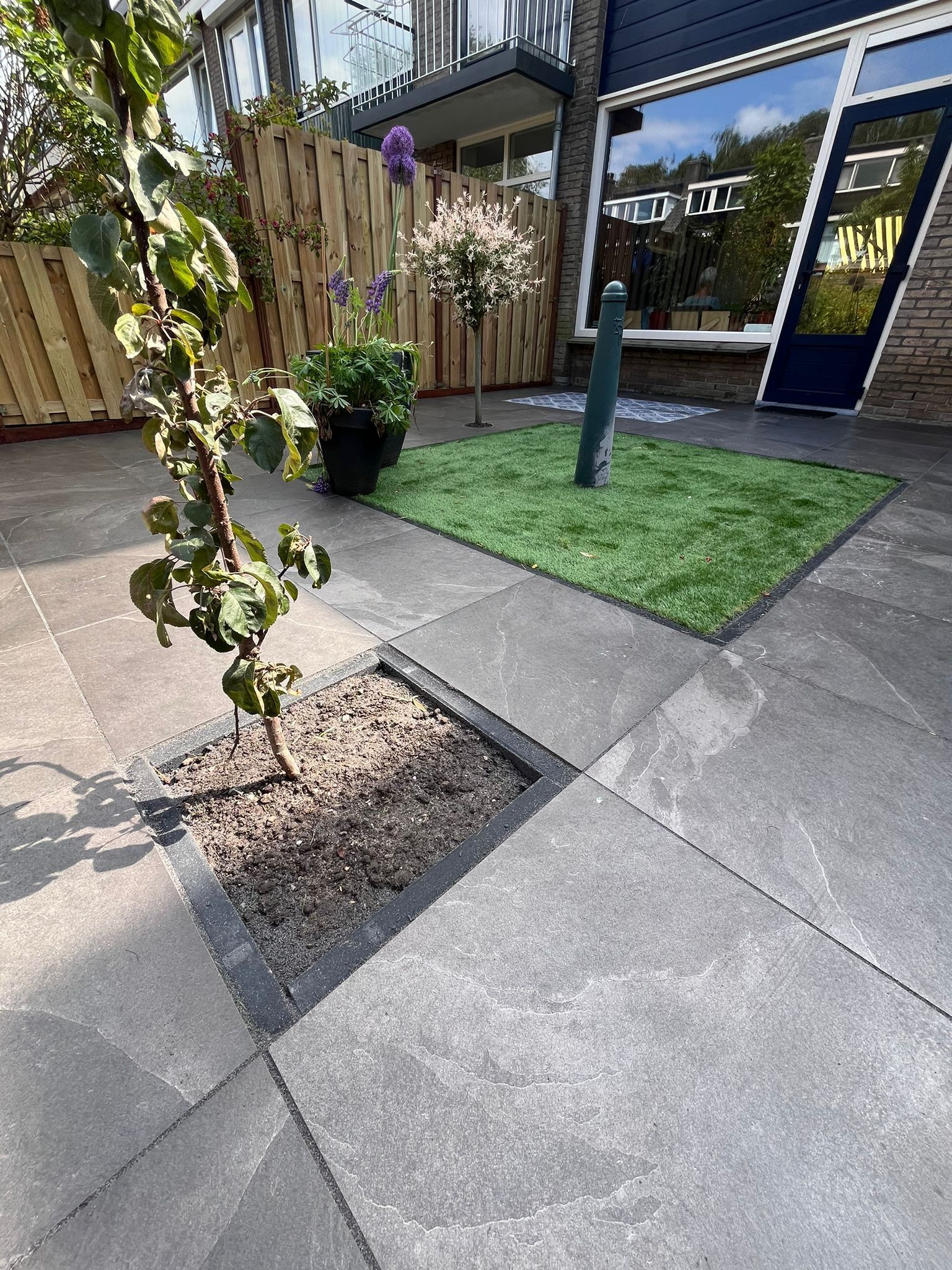 Terras met vierkante bestrating, een klein boompje in een plantenbak en een stukje kunstgras.