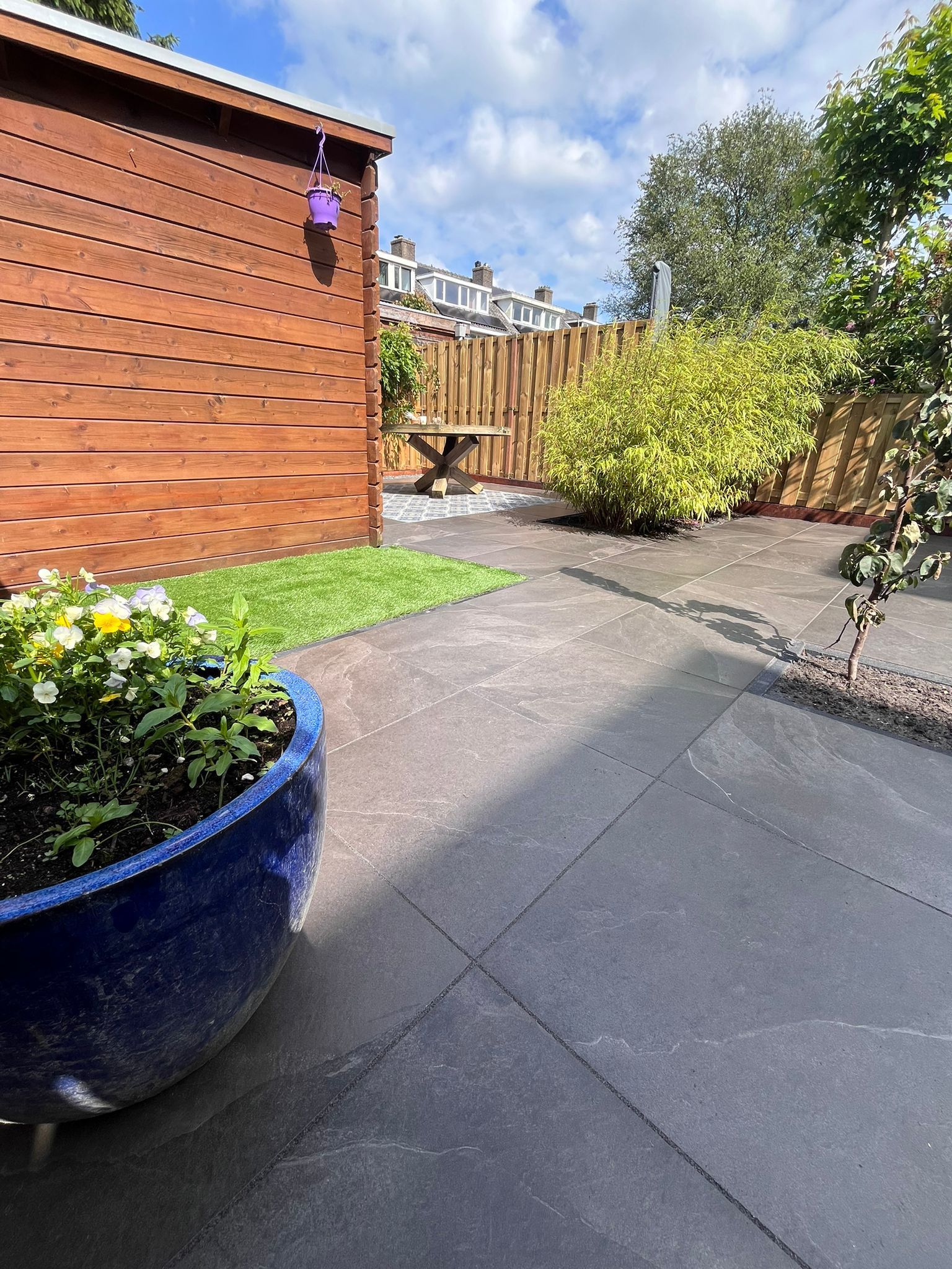 Een terras in de achtertuin met donkergrijze bestrating en een blauwe plantenbak. Houten schuttingen en groen omringen het gebied.