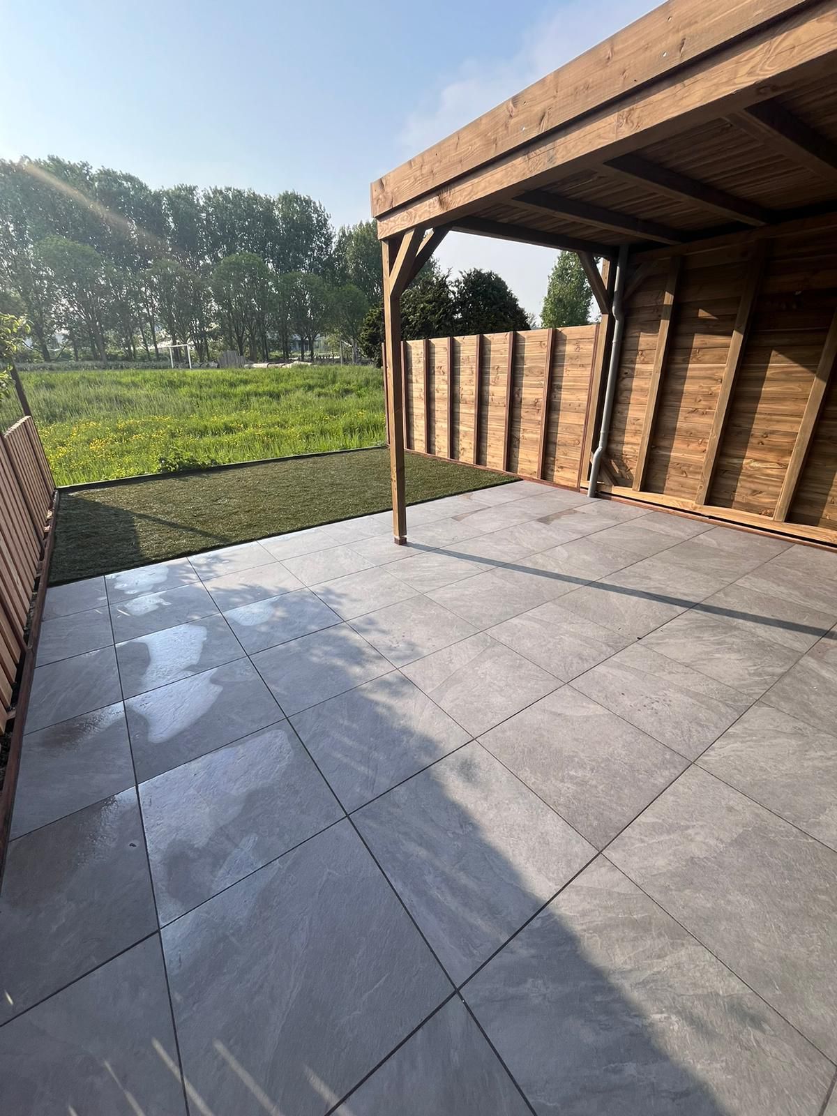 Terras met grijze tegelvloer, houten pergola en schutting; groen gazon op de achtergrond.