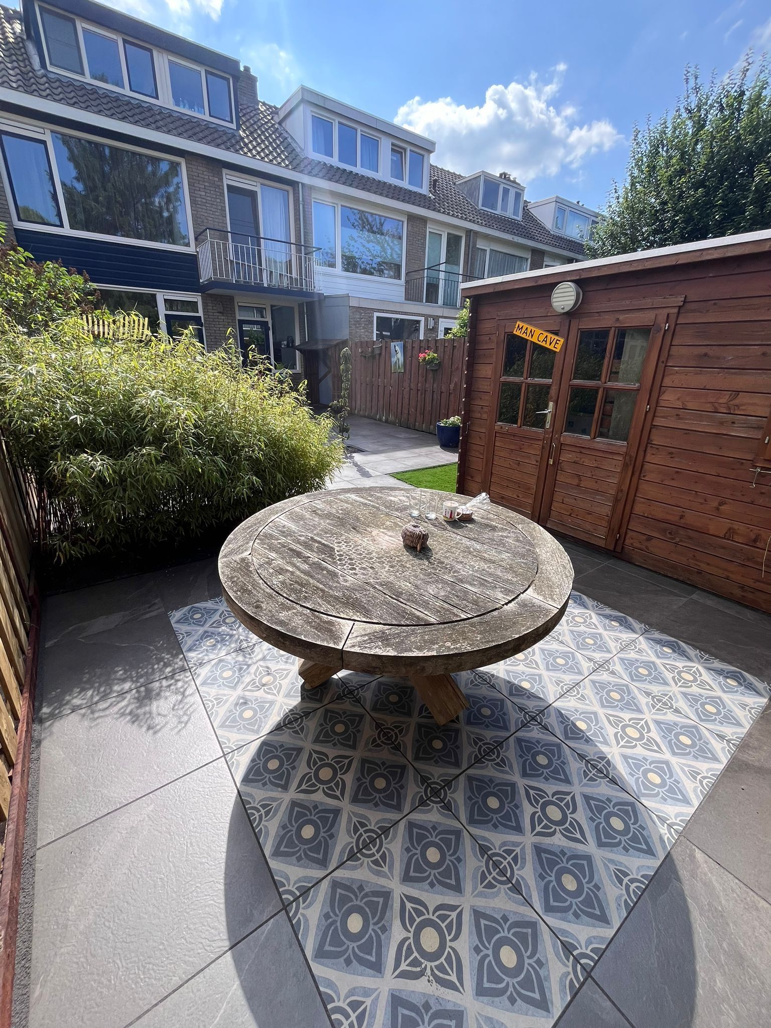 Terras met ronde tafel op decoratieve tegels, omgeven door een schuur, een hek en huizen onder een zonnige hemel.