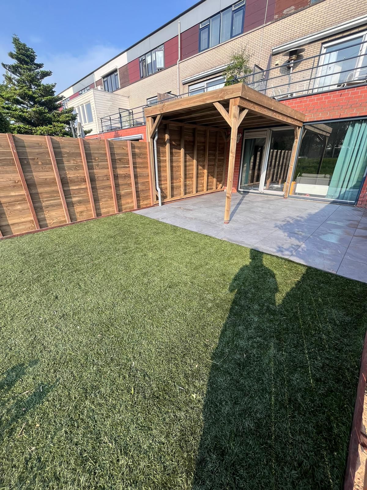 Achtertuin met kunstgras, houten schutting, pergola en huis.