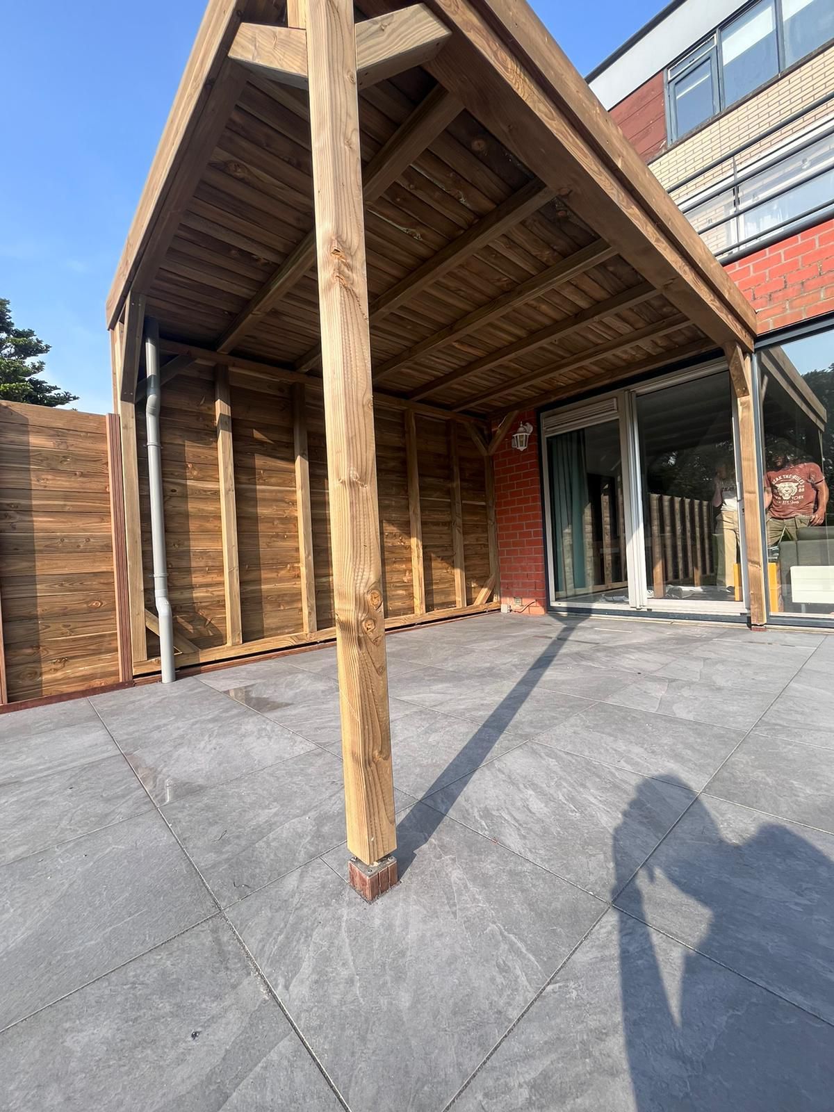 Houten terrasoverkapping met steunpaal op een grijs betegeld terras, vlakbij een gebouw met glazen deuren en ramen.