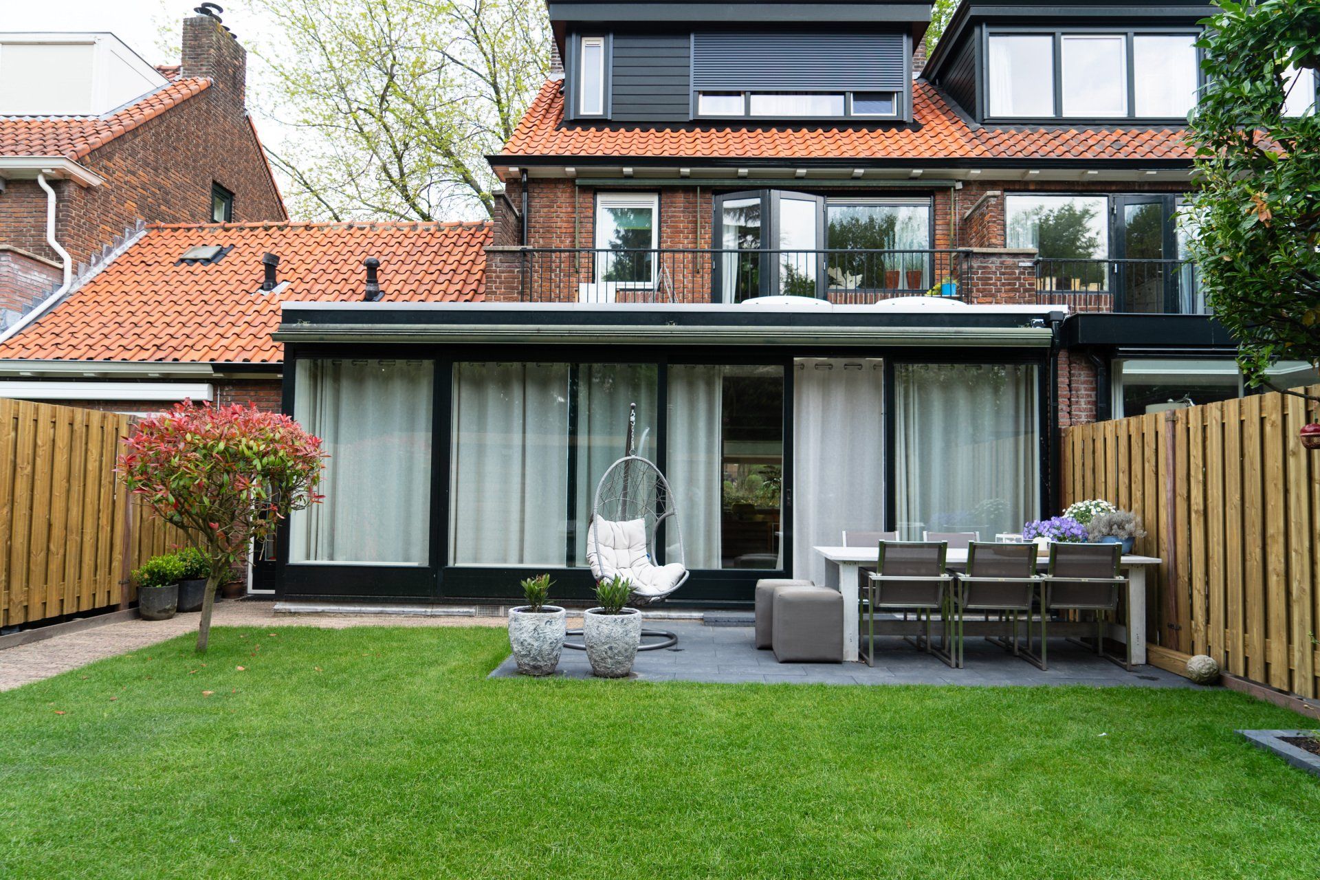 Achtertuin met gras, terras en een modern huis met glazen deuren en een rood pannendak.