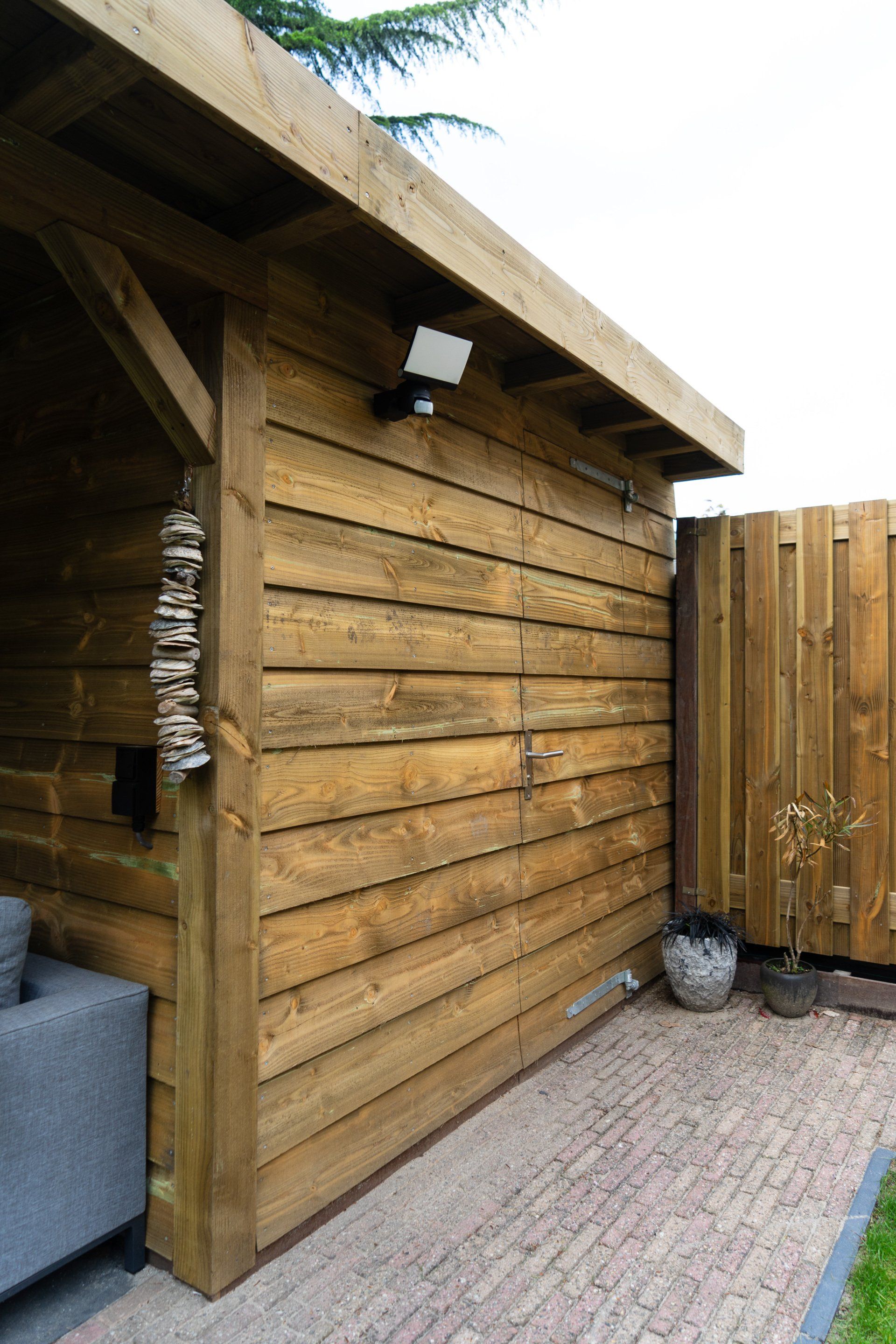Houten buitenconstructie met horizontale zijwanden en een veiligheidslamp. Bakstenen terras en houten schutting op de achtergrond.