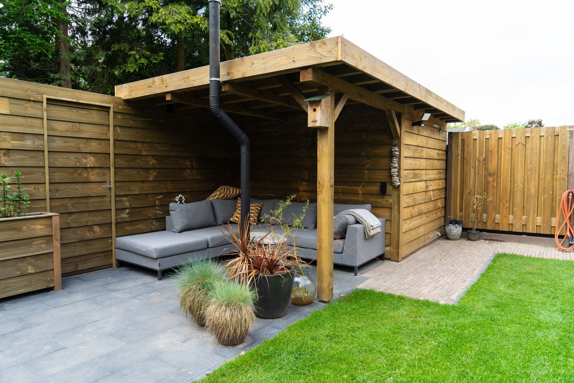 Houten terrasoverkapping met grijs meubilair, op een grijs stenen terras, grenzend aan een groen gazon.