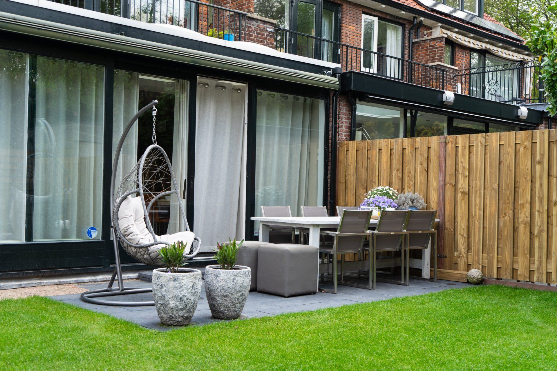 Terras in de achtertuin met hangstoel, eettafel en houten schutting. Omgeven door groen gras.