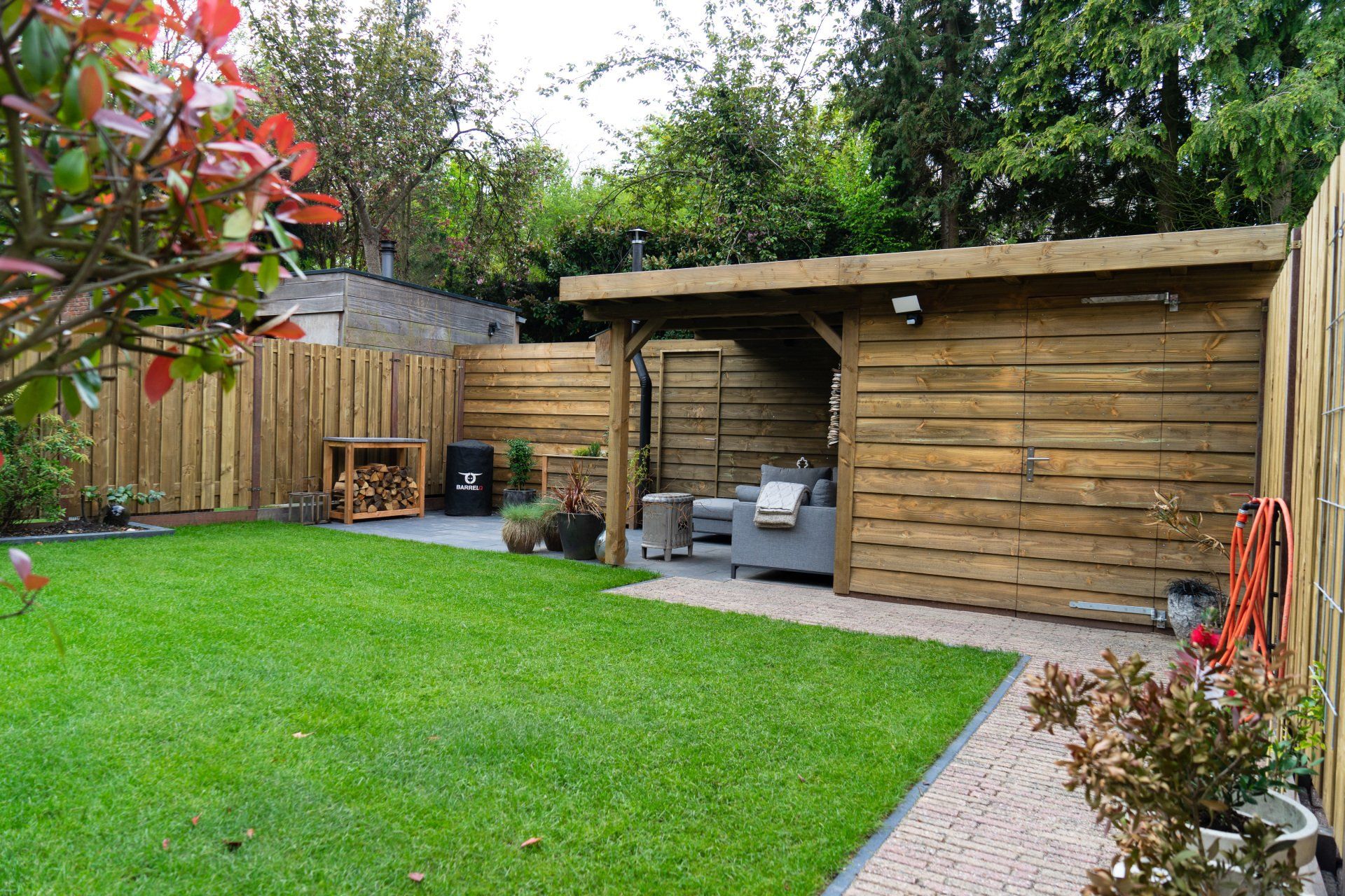 Achtertuin met groen gras, houten constructies om te barbecueën en spullen op te bergen, en een stenen pad.