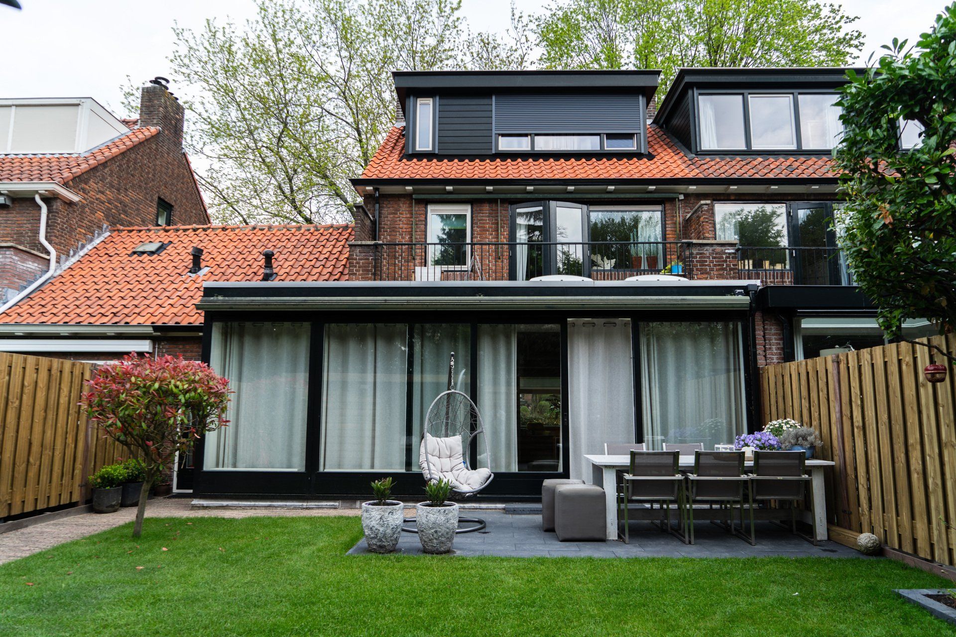 Achtertuin met huis, terras en gazon. Het huis heeft een rood pannendak, zwarte kozijnen en grote ramen. Buitenmeubilair en hangstoel.