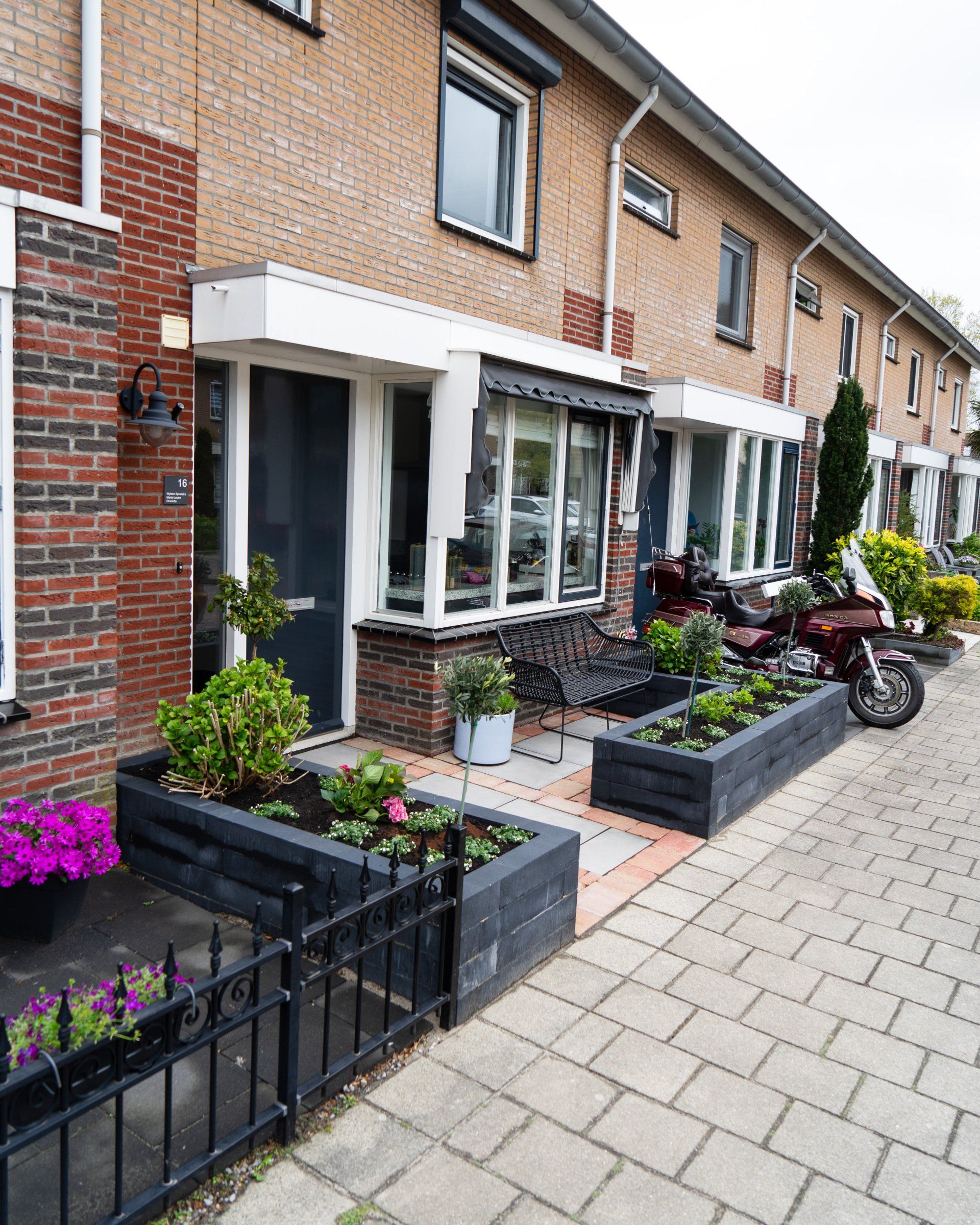 Een rij bakstenen rijtjeshuizen met bloembakken, planten, een motorfiets en een zwart hek.