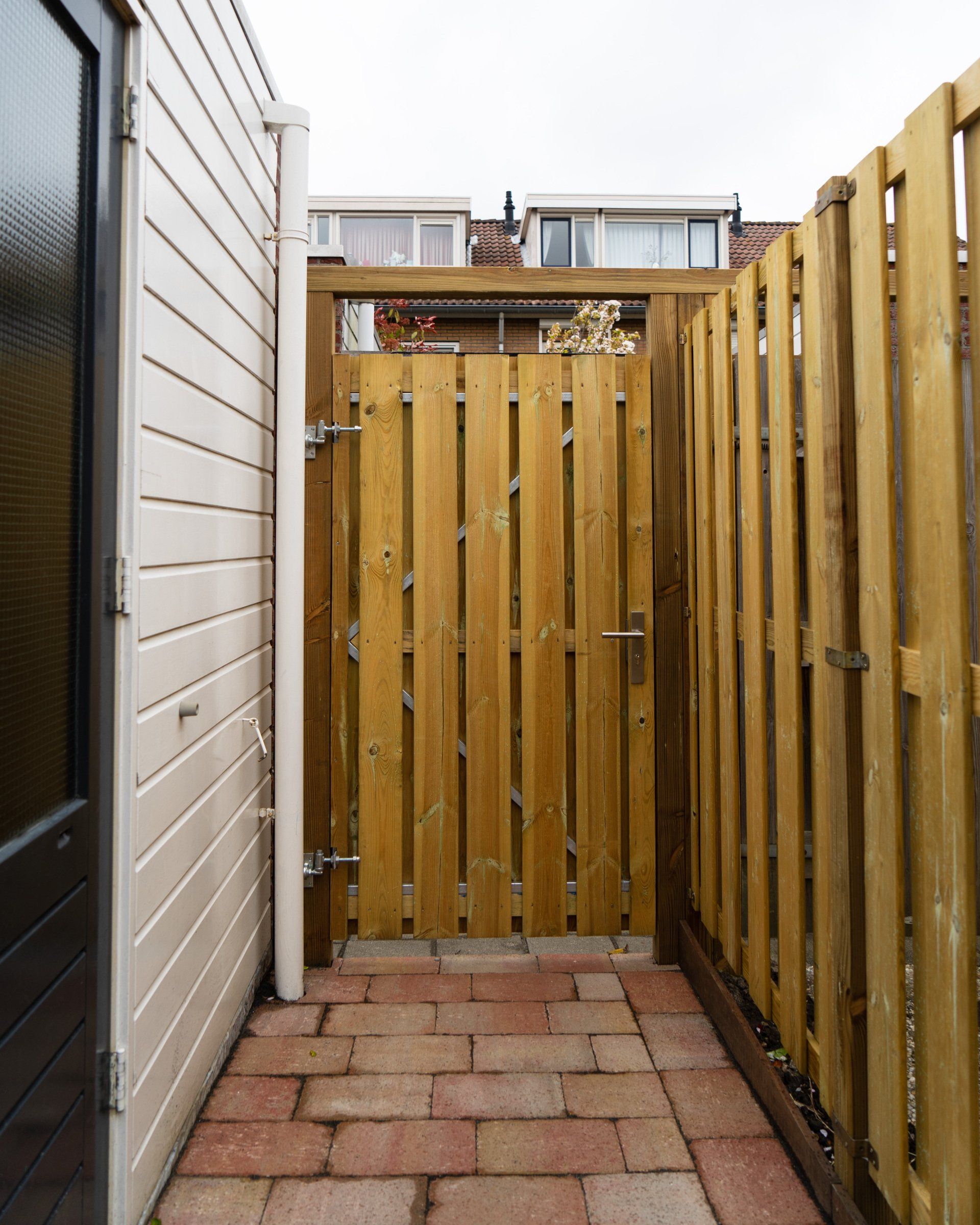 Houten poort in een smal, met bakstenen bestrate steegje tussen een wit gebouw en een houten hek.