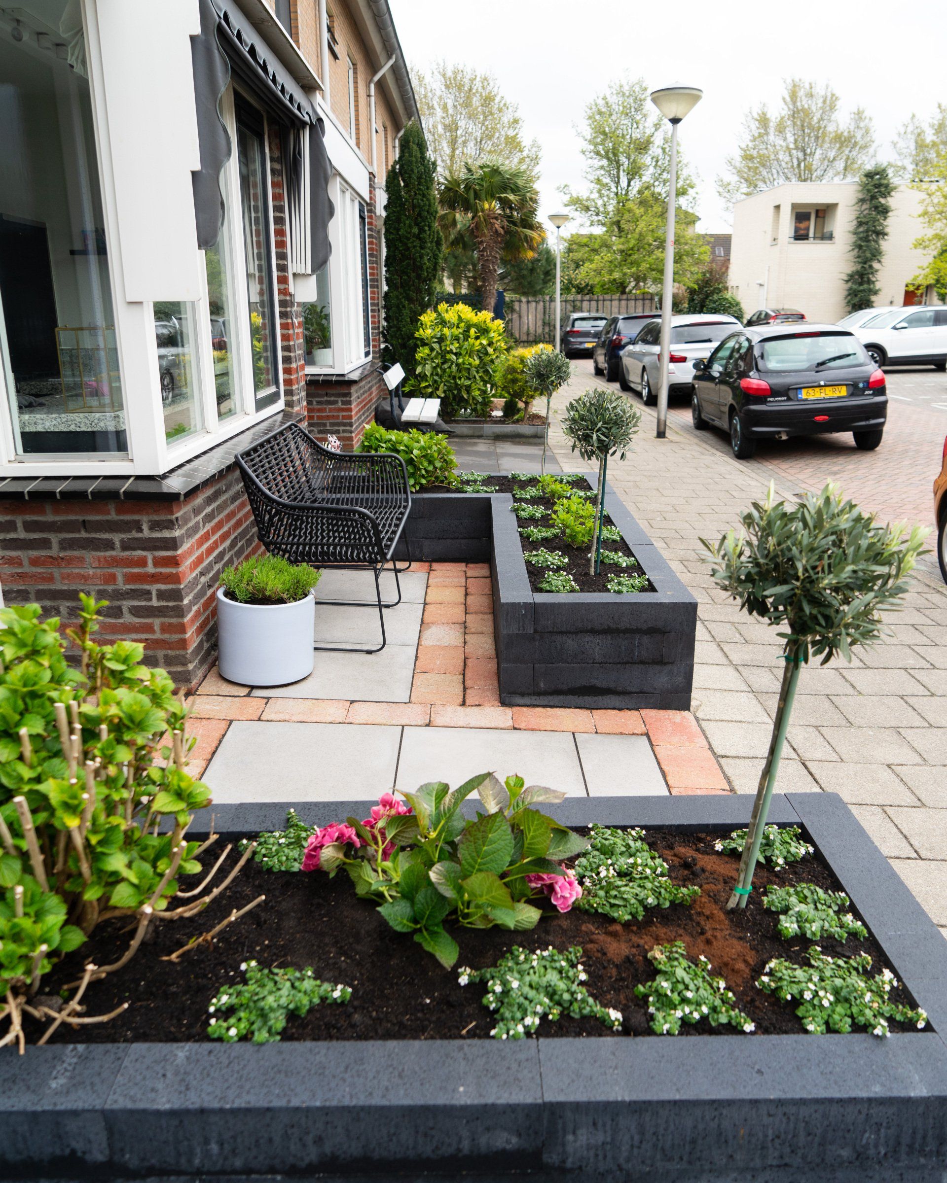 Voortuin met donkere, verhoogde bloembedden, bloemen, een pad van bakstenen en stenen, een zwarte bank en geparkeerde auto's.
