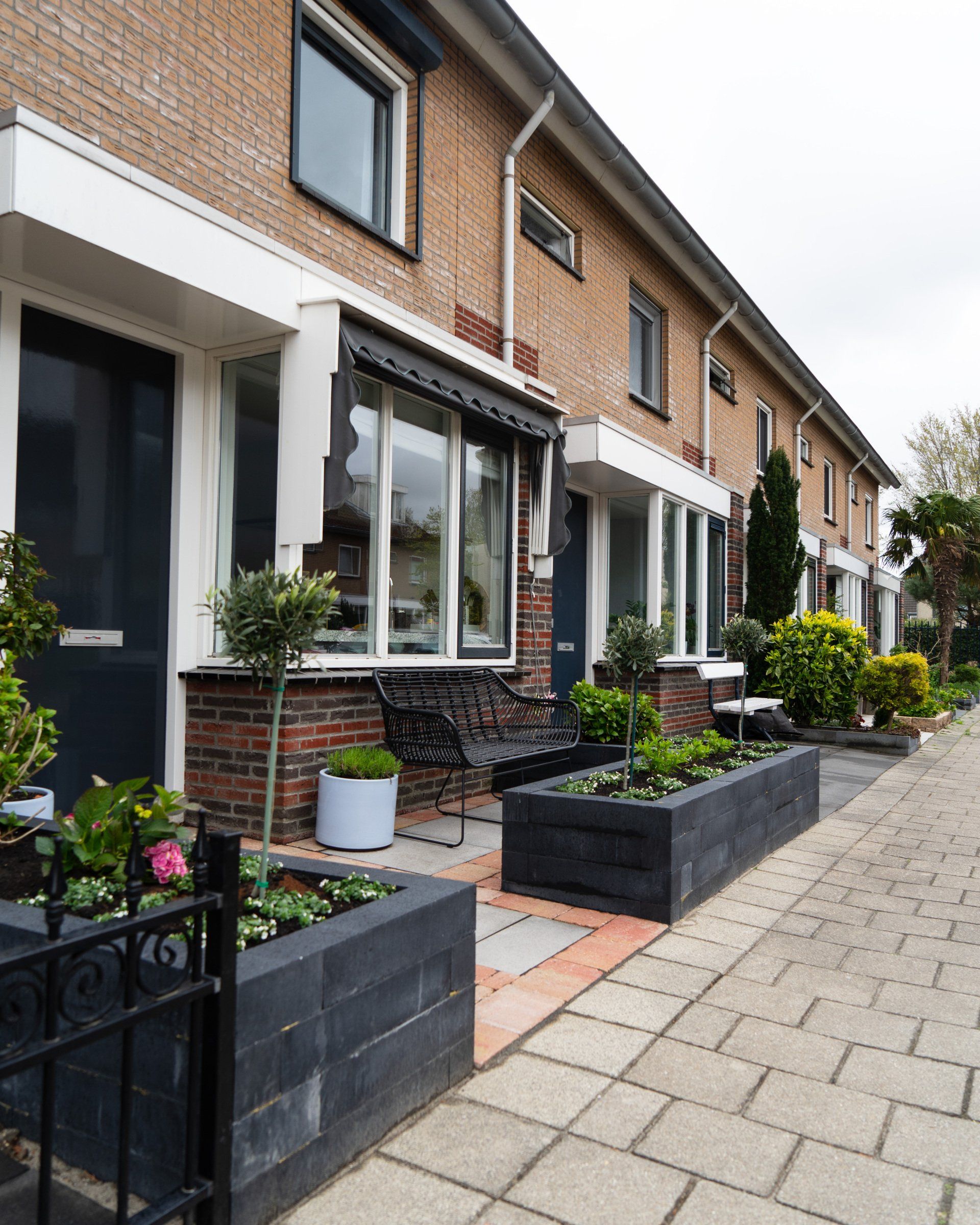 Een rij bakstenen herenhuizen met donkere deuren, plantenbakken en een trottoir.