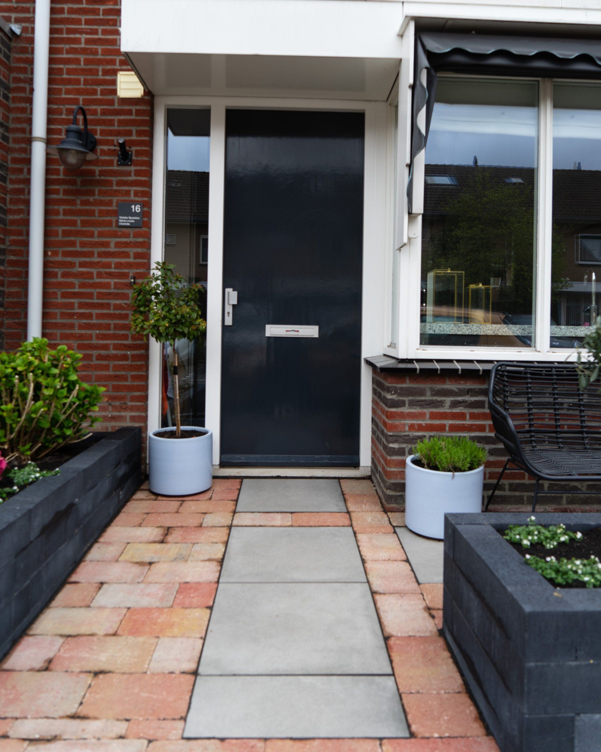 Vooringang met een donkere deur, bakstenen gevel en een bestraat pad met plantenbakken.