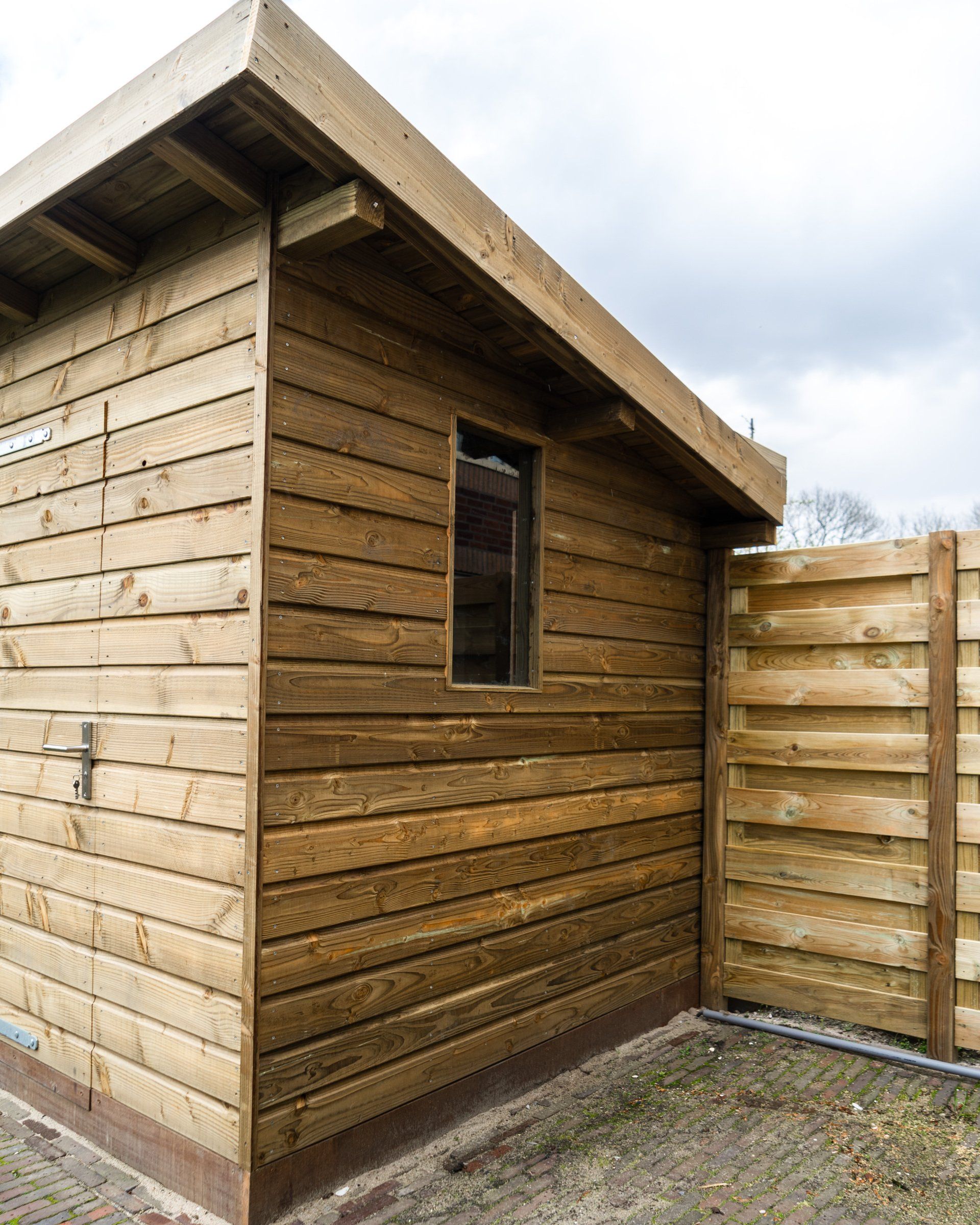Houten schuur met één raam, naast een houten schutting.