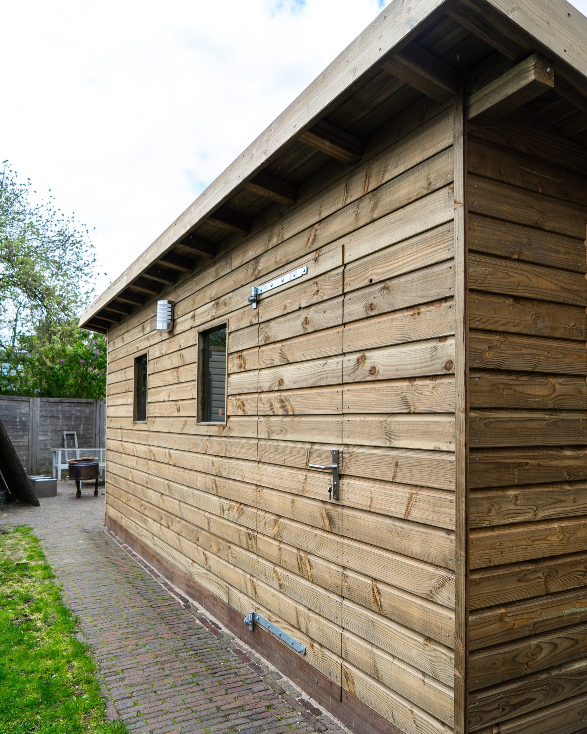 Houten schuur met horizontale planken als gevelbekleding, ramen en een deur, gelegen buiten op een bestraat terrein met een groen pad.