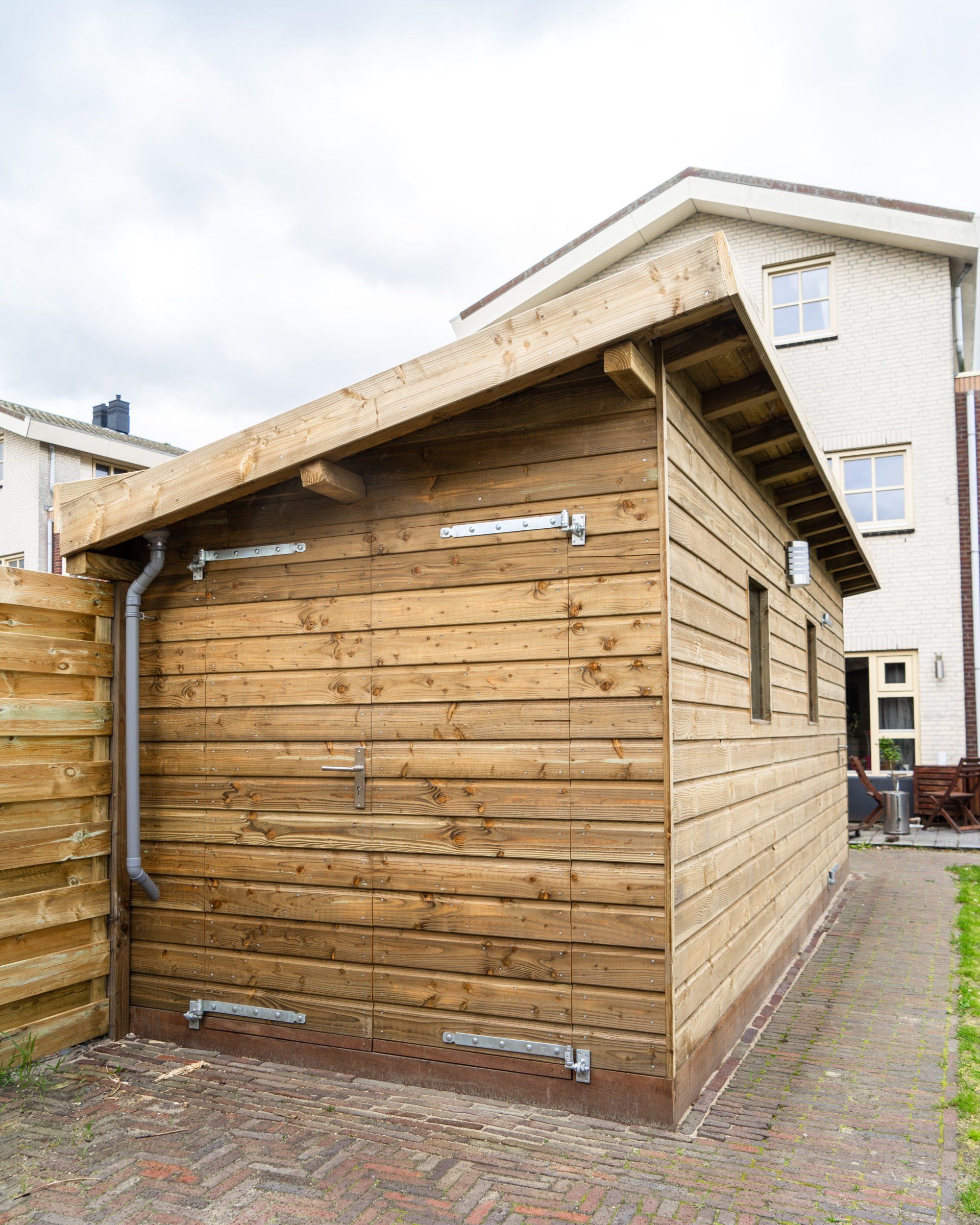 Houten schuur met horizontale planken als gevelbekleding, goten en een schuin dak, naast een hek en een bijgebouw.