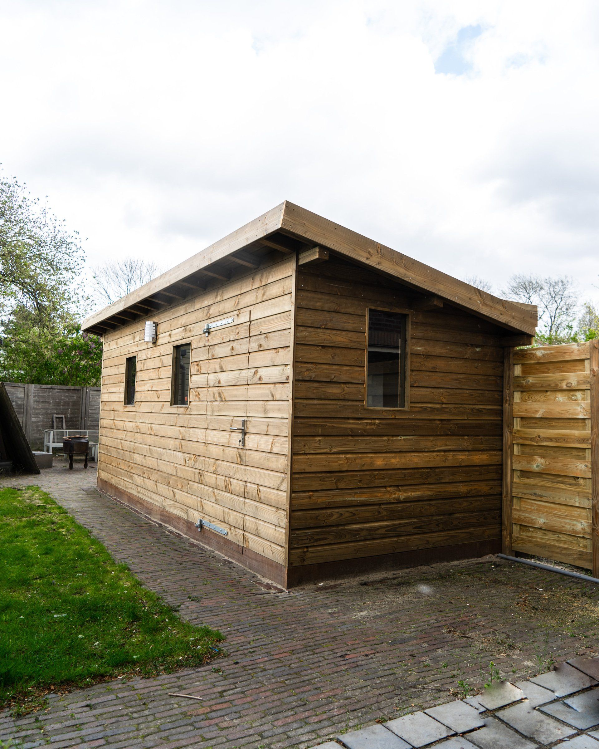 Houten schuur met drie ramen, bruine gevelbekleding, naast een bestraat gedeelte en gras.