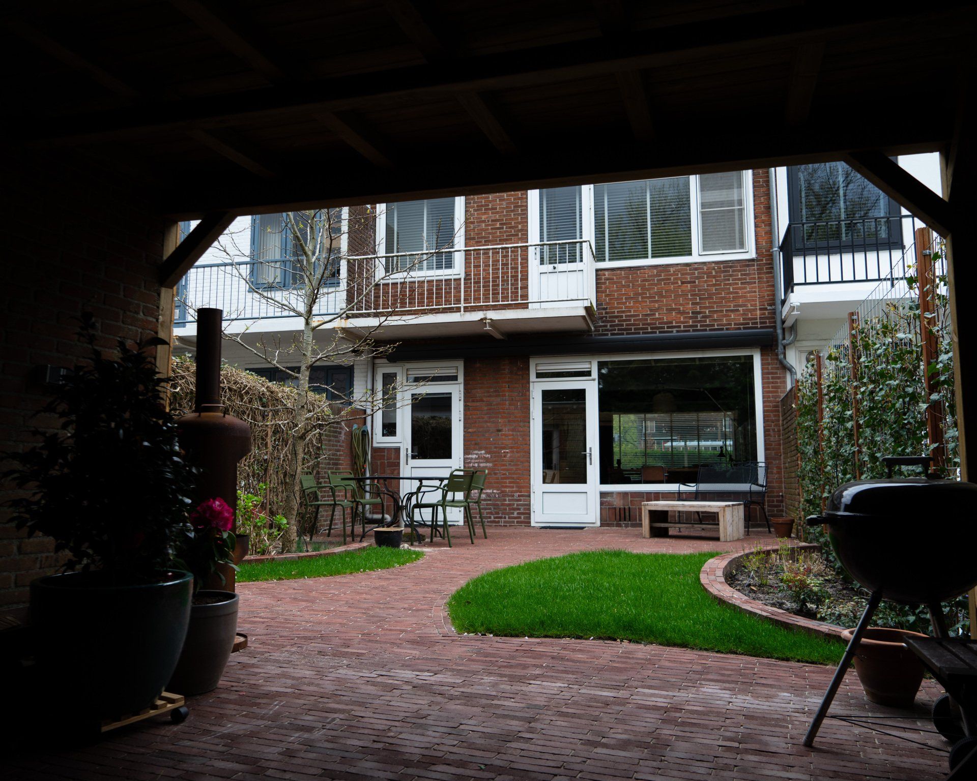 Een stenen terras met groen gras leidt naar een stenen gebouw met witte ramen, dat te zien is vanaf een overdekte ruimte.