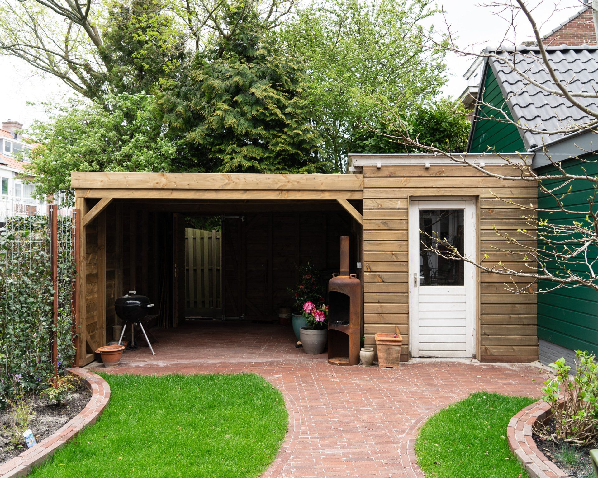Houten carport met een stenen terras, een deur en een barbecue in een achtertuin.