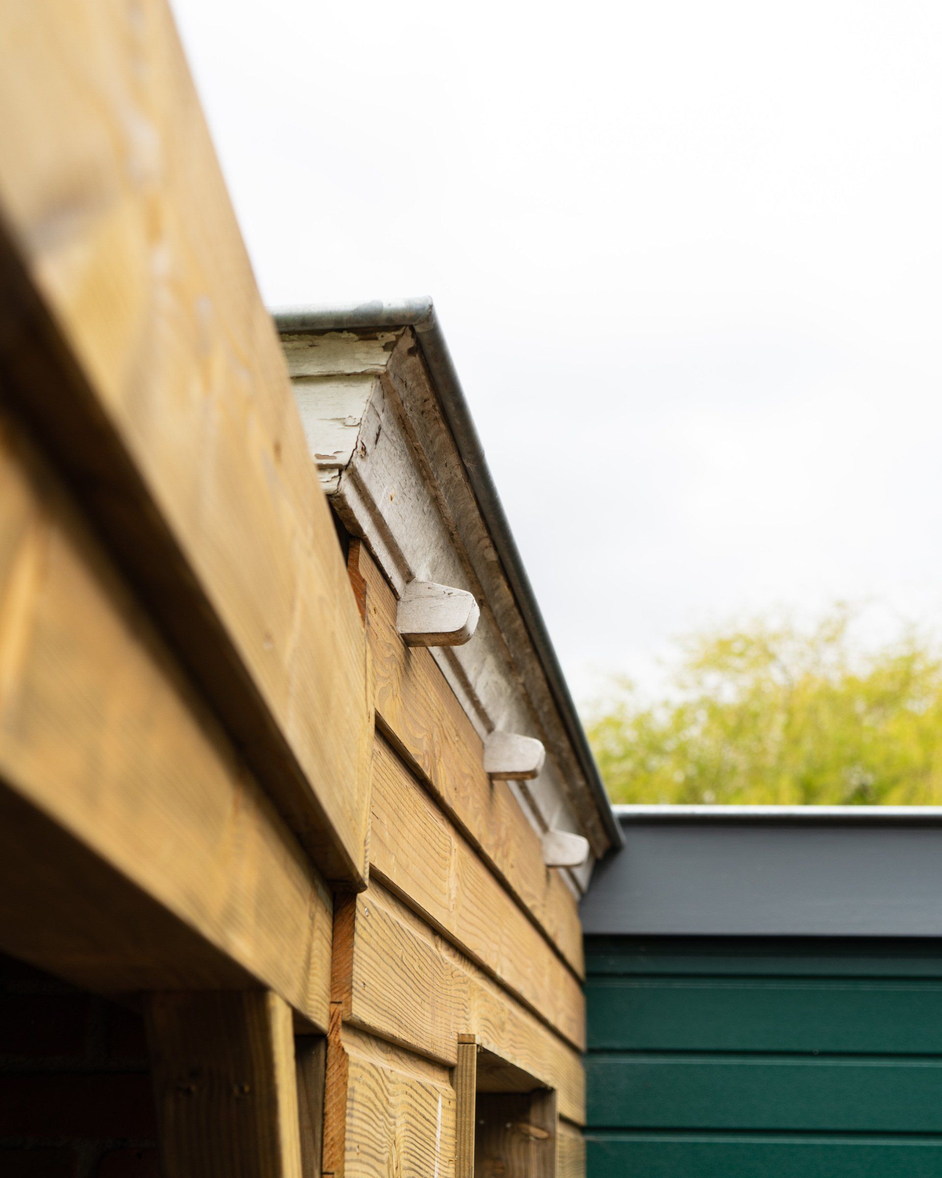 De buitenkant van een houten schuur met witte beugels die de dakrand ondersteunen; een groene constructie is zichtbaar.