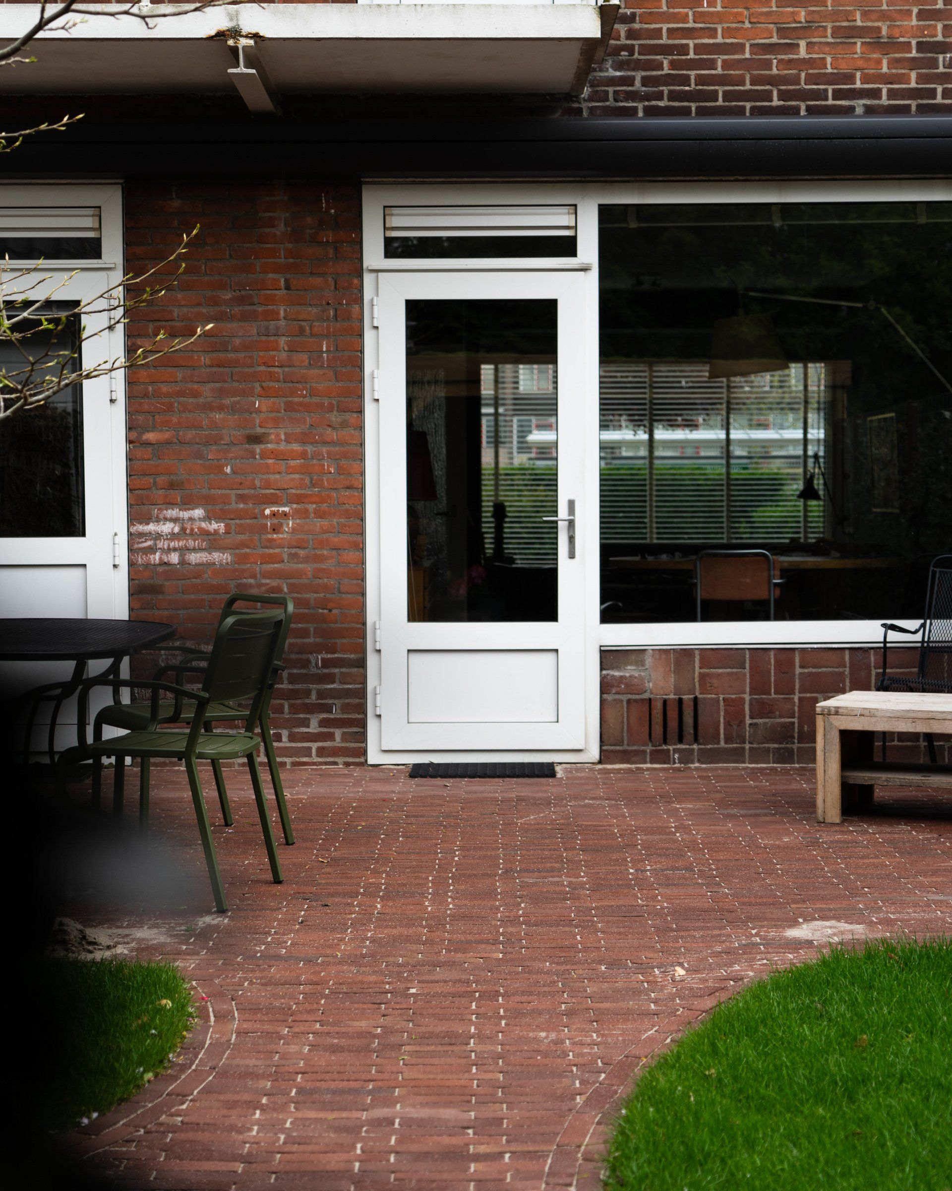 Terras van rode bakstenen met een witte deur en raam, groene stoelen en een gazon.