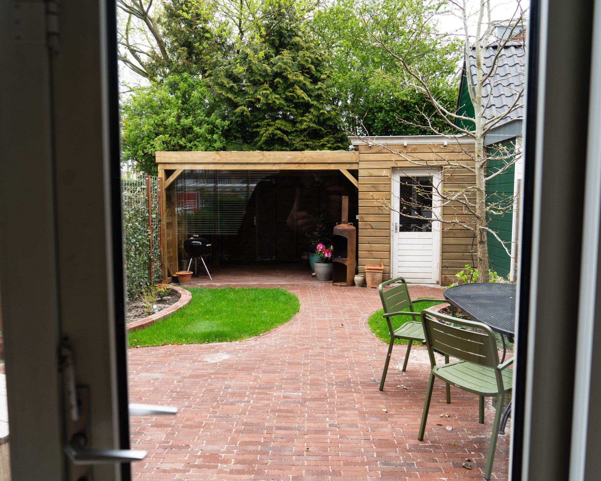 Uitzicht op een achtertuin met een stenen terras, een klein gazon, een houten afdak, groene stoelen en een kleine witte deur.