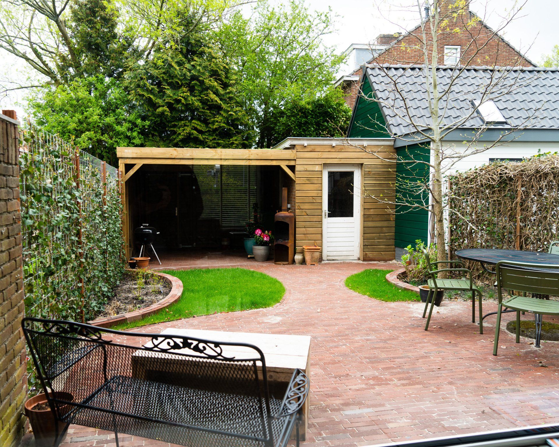 Een stenen terras met een houten schuur, een bankje en groen gras.