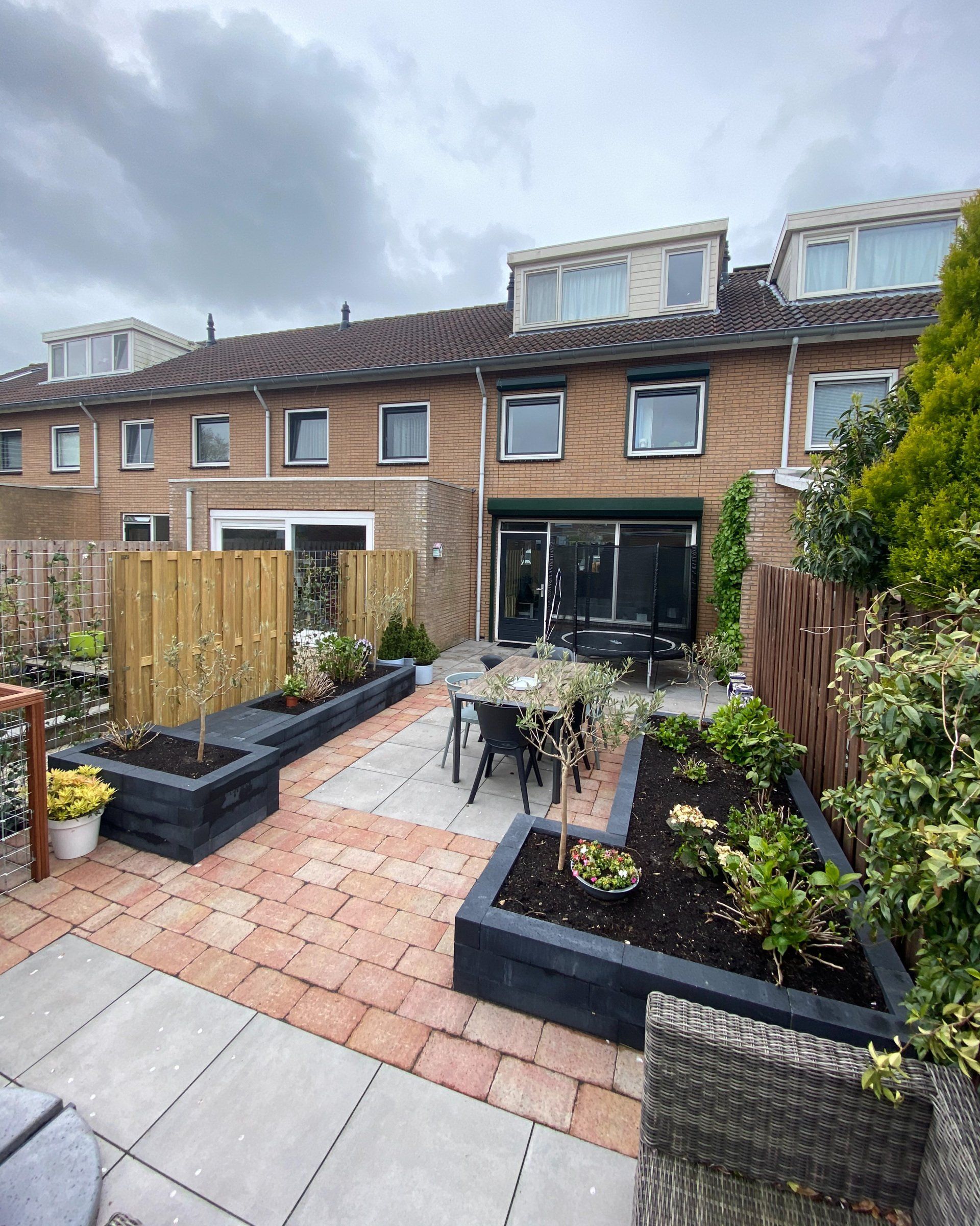 Terras in de achtertuin met verhoogde bloembedden en een eethoek, gelegen tegen een bakstenen huis.