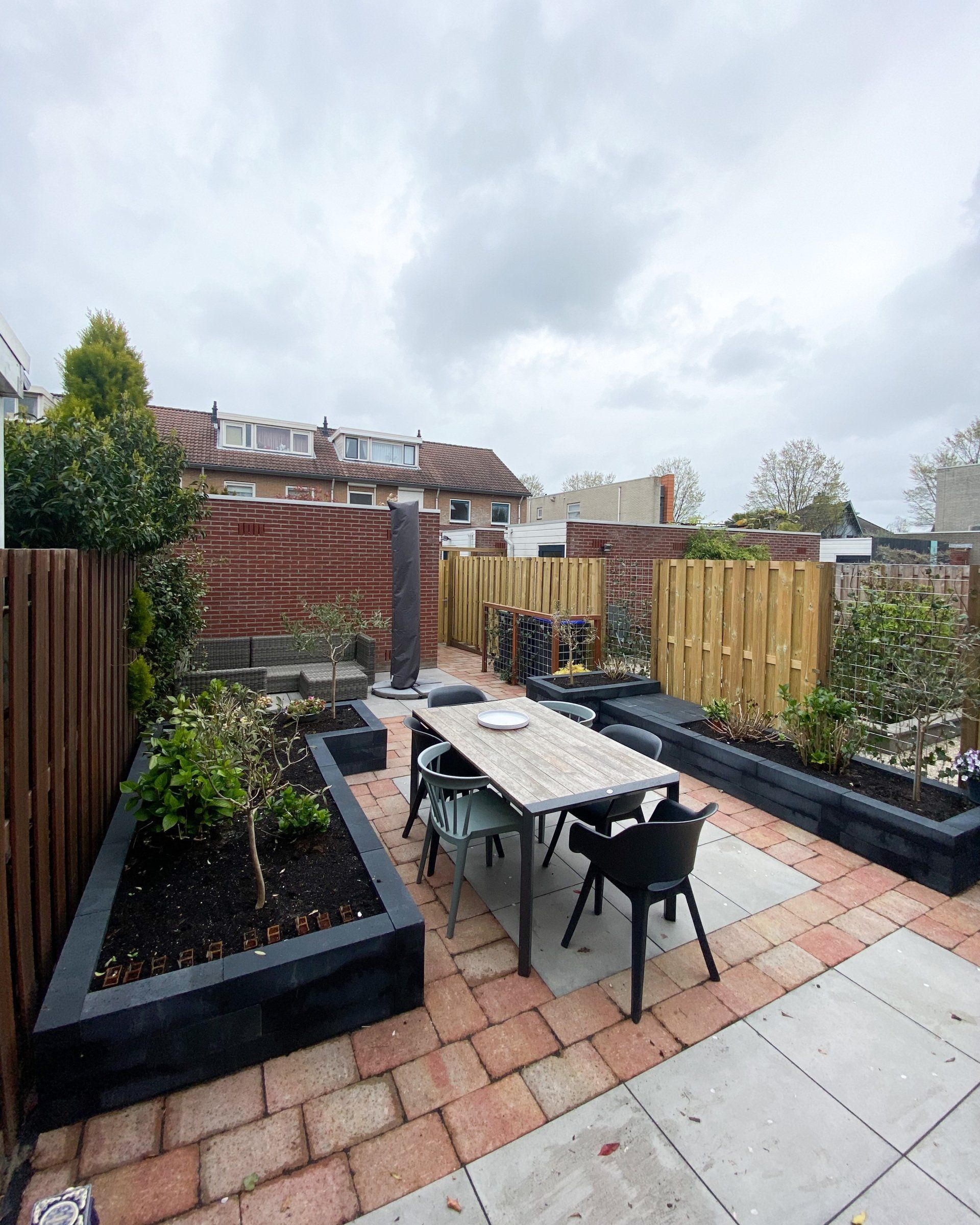 Terras buiten met bakstenen bestrating, een houten tafel, donkere stoelen en verhoogde plantenbakken. Bewolkte lucht.