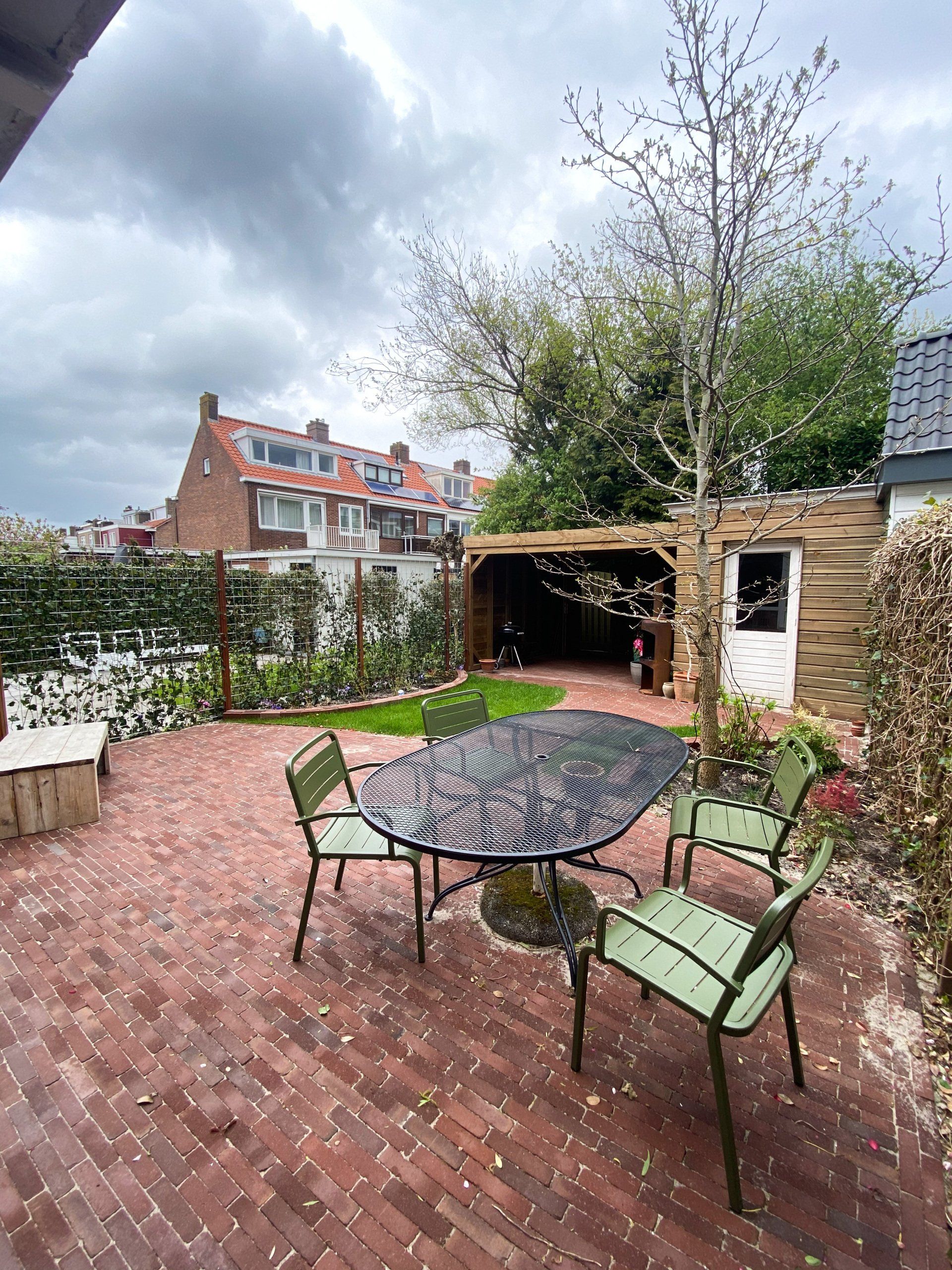 Een stenen terras met groene stoelen en een tafel, een pergola en omringende begroeiing onder een bewolkte hemel.