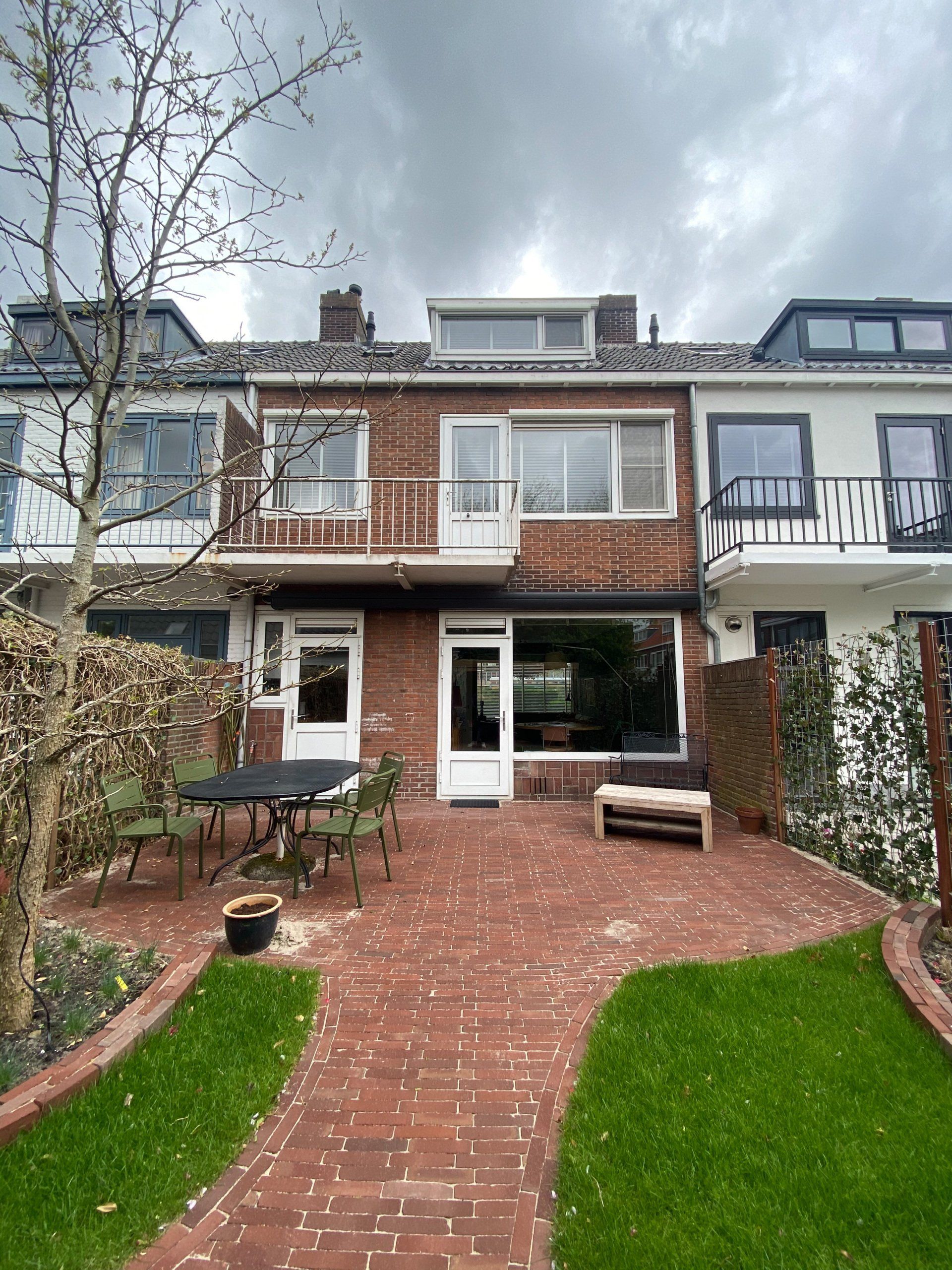 Rood bakstenen huis met terras, groen gazon, tafel en stoelen. Bewolkte lucht.