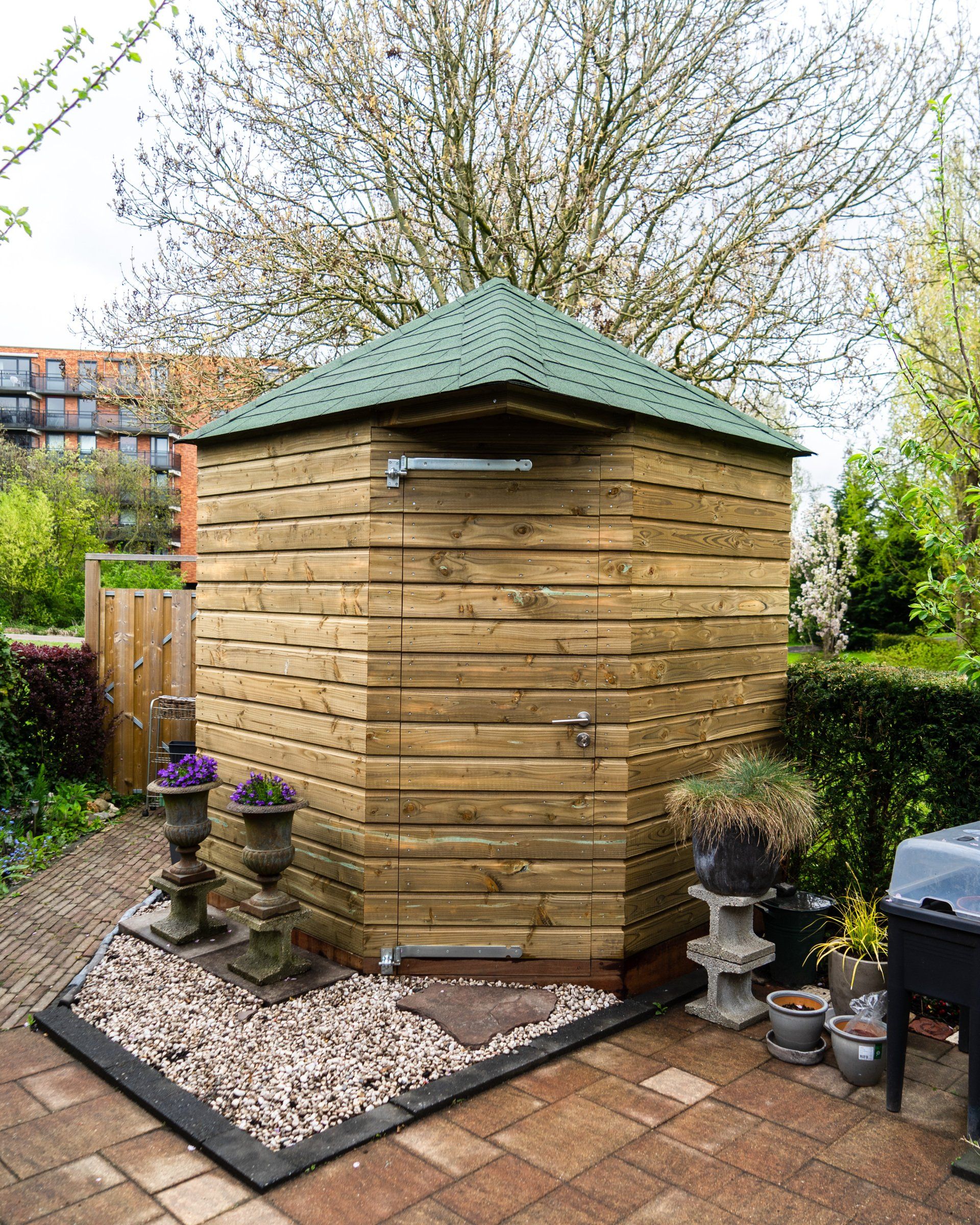 Houten zeshoekige schuur met groen dak in een achtertuin, omgeven door planten en een stenen terras.
