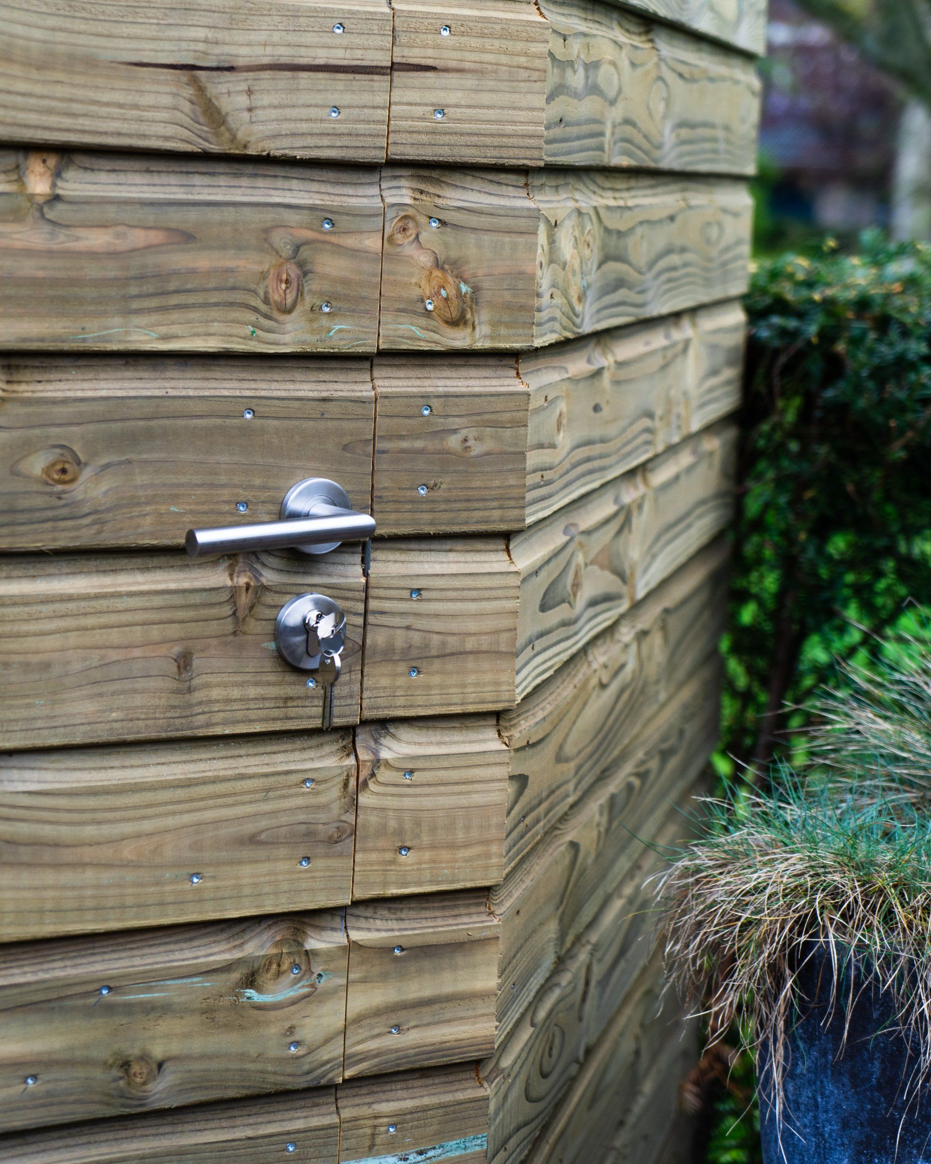 Houten hoekhuisje met deur en metalen handgreep, gelegen in een tuin met groen.