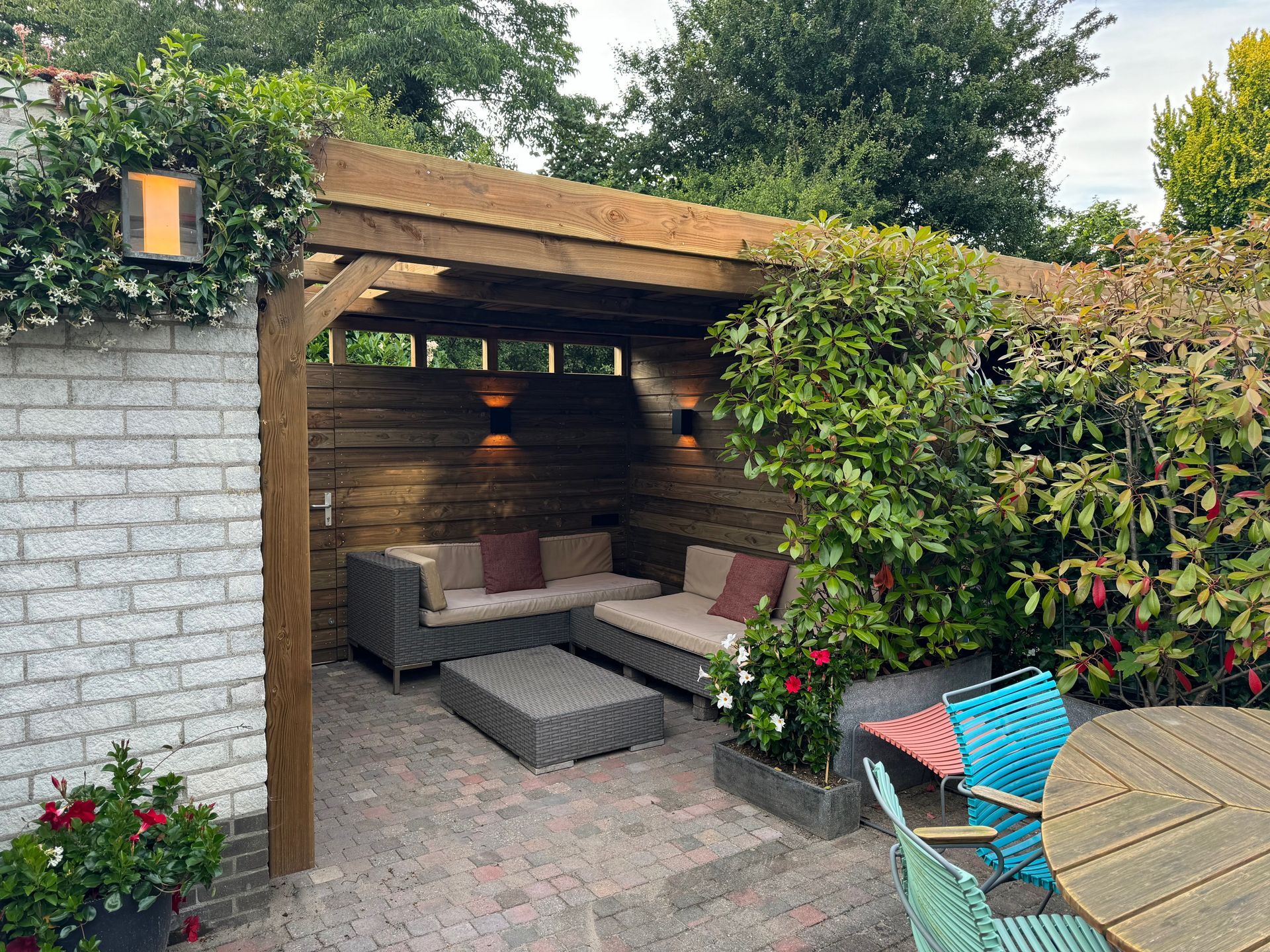 Gezellige zithoek buiten met houten pergola, grijze bank en weelderig groen.