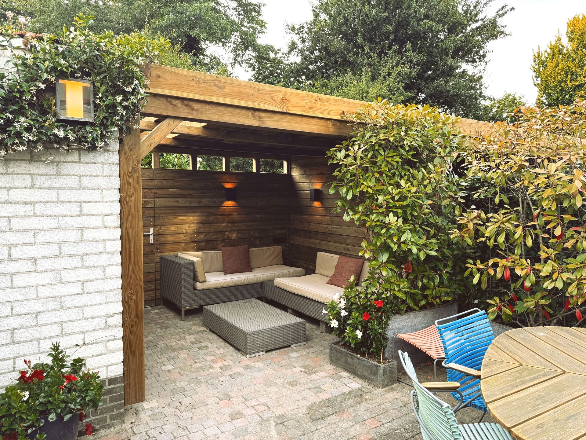 Buiten zitgedeelte met houten pergola, bank en tafel. De ruimte is omgeven door groene begroeiing.