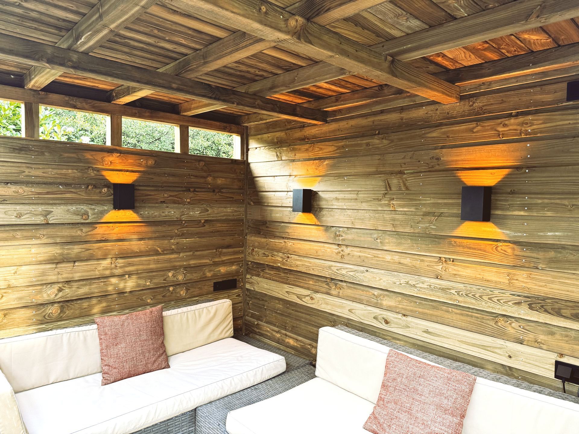 Houten prieel met zitgelegenheid, verlicht door warme wandlampen.