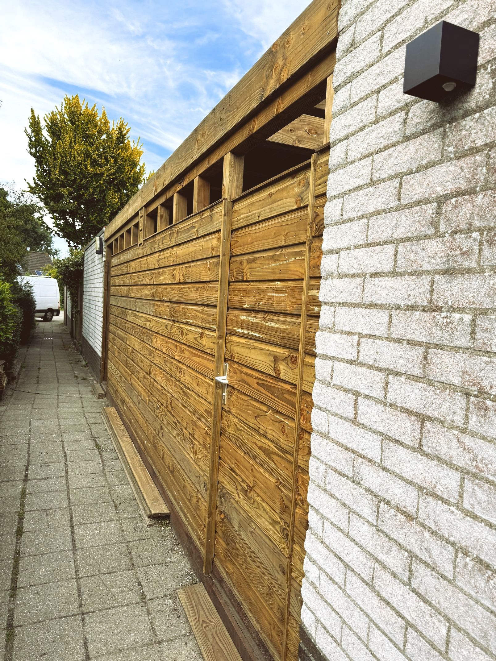 Een houten schutting naast een witte bakstenen muur en een geplaveid pad onder een blauwe hemel.