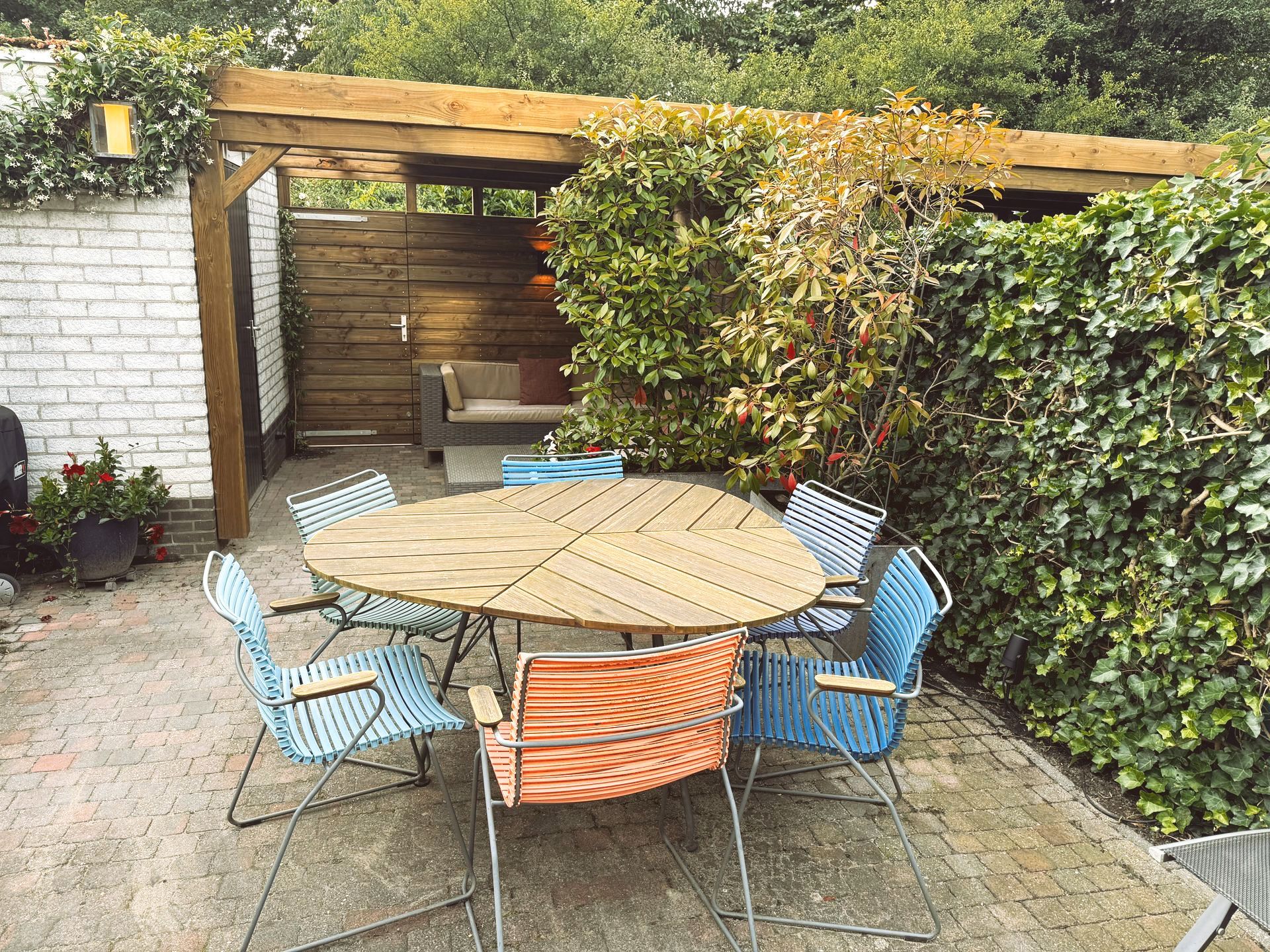 Buitenpatio met houten pergola, tafel en kleurrijke stoelen, omgeven door groen.