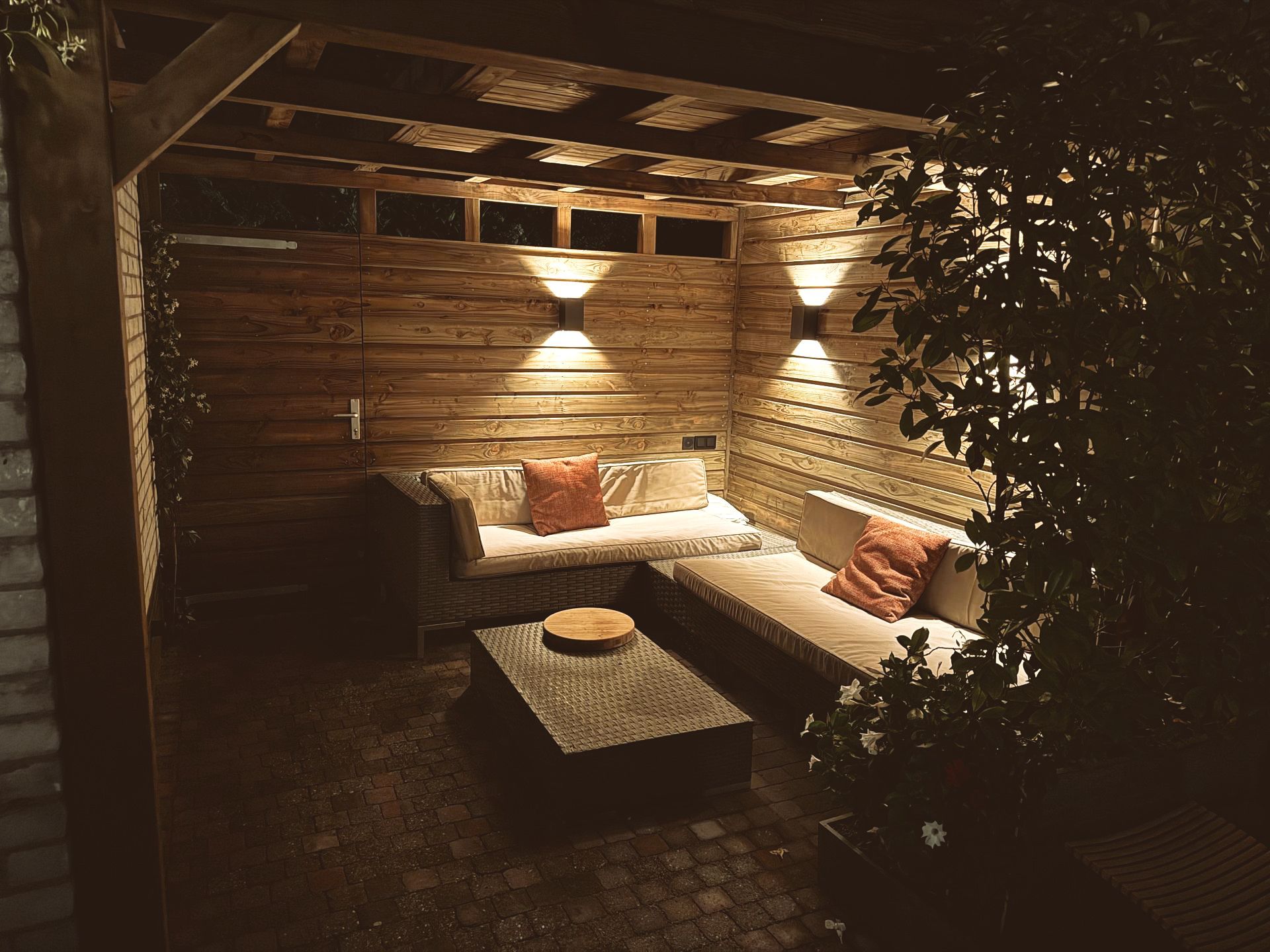 Gezellige loungeplek buiten 's avonds met bank, kussens, salontafel en wandlampen.