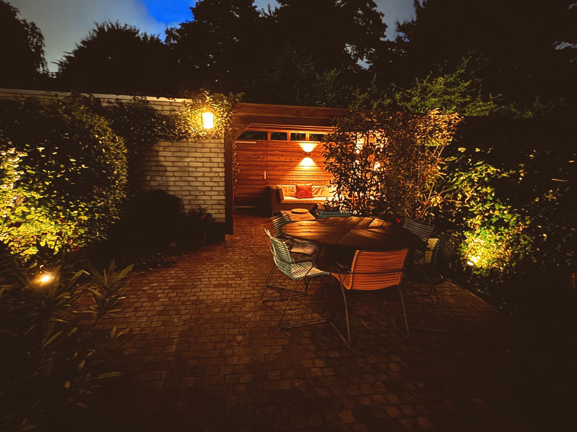 Avondtafereel: verlichte eethoek in de buitenlucht met bakstenen terras, houten tafel en weelderig groen.
