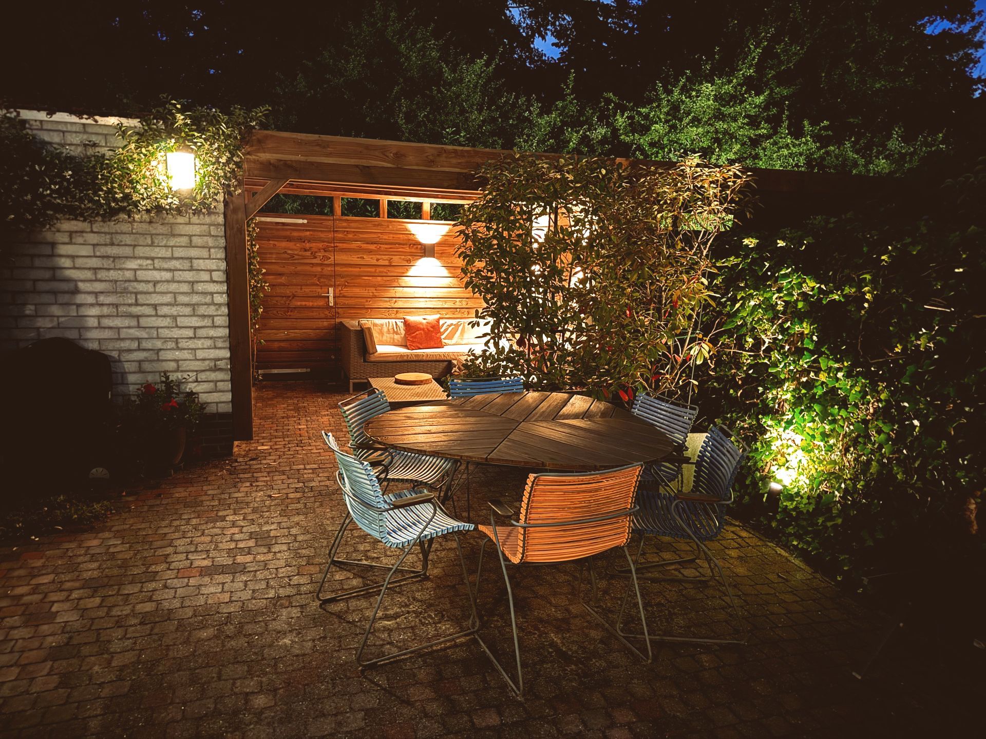 Buiten eetgedeelte 's avonds, verlicht door lichtslingers en plafondlampen. Houten tafel en stoelen.