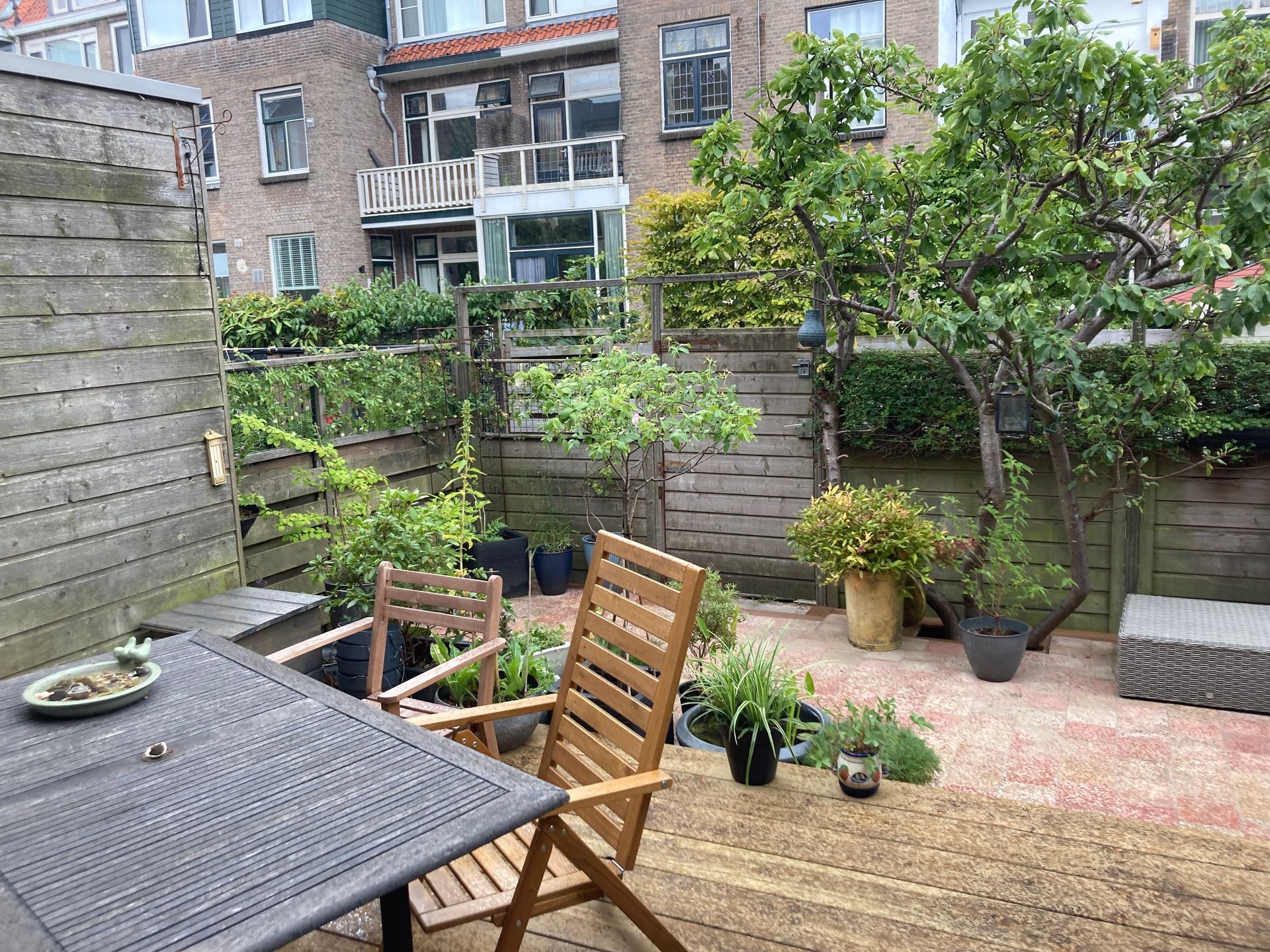 Terras in de achtertuin met tafel, stoelen, planten en een houten schutting. Op de achtergrond zijn bakstenen gebouwen te zien.