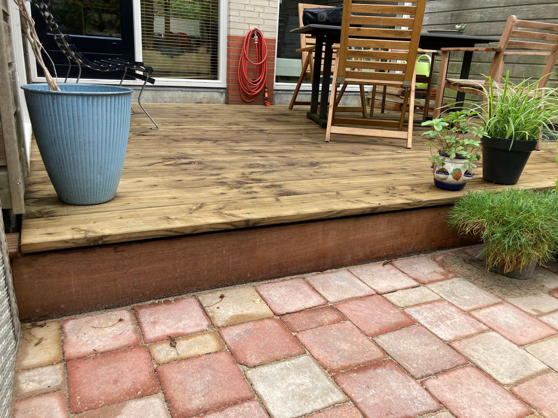 Houten terras met een blauwe plantenbak, tuinmeubilair en bakstenen bestrating.