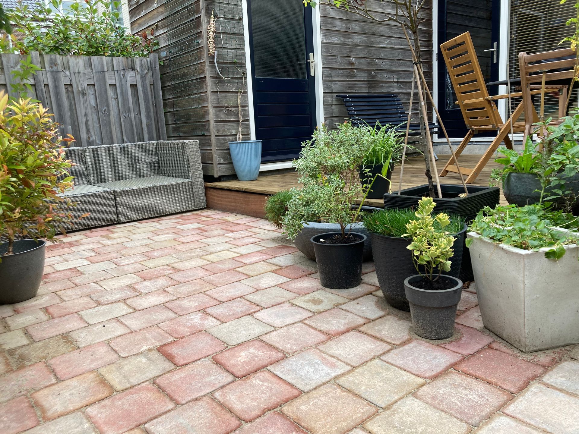 Bakstenen terras met potplanten, houten schuur, schutting en zitgelegenheid.