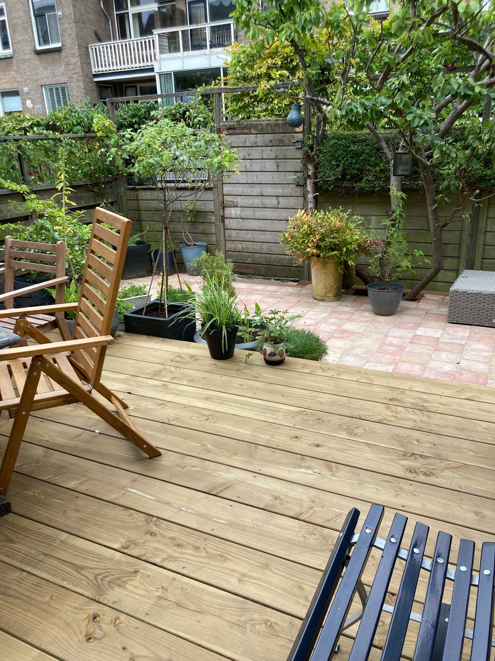 Houten terras met stoelen, planten en een stenen patio in een tuin.