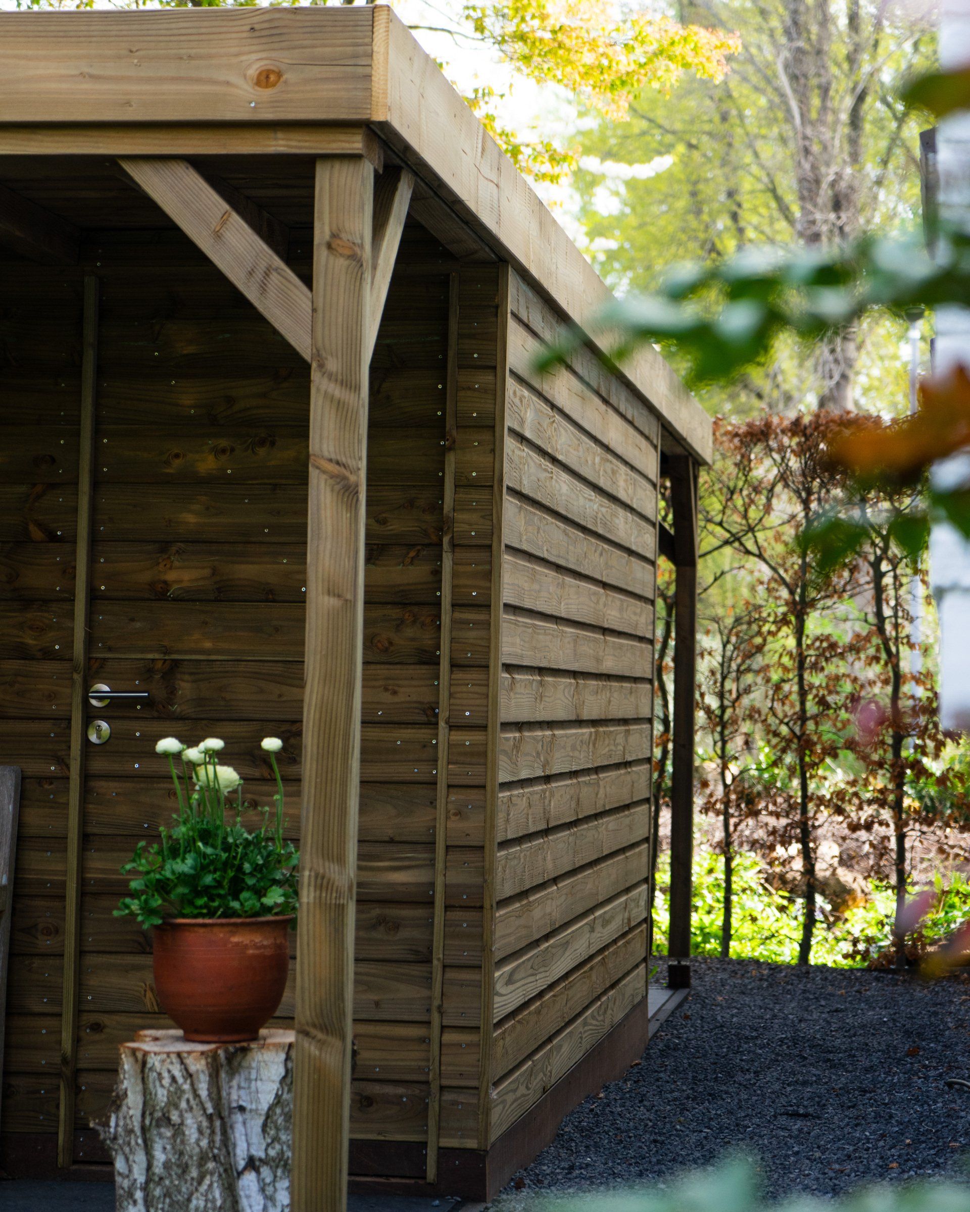 Houten schuur met schuin dak, donkerbruine gevelbekleding, een bloempot en een boomstronk in een tuinomgeving.