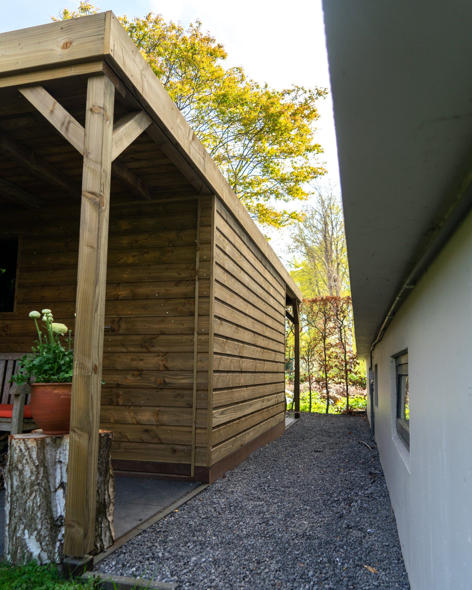 Houten schuur met donkere gevelbekleding, naast een wit gebouw. ​​Grindpad ertussen.